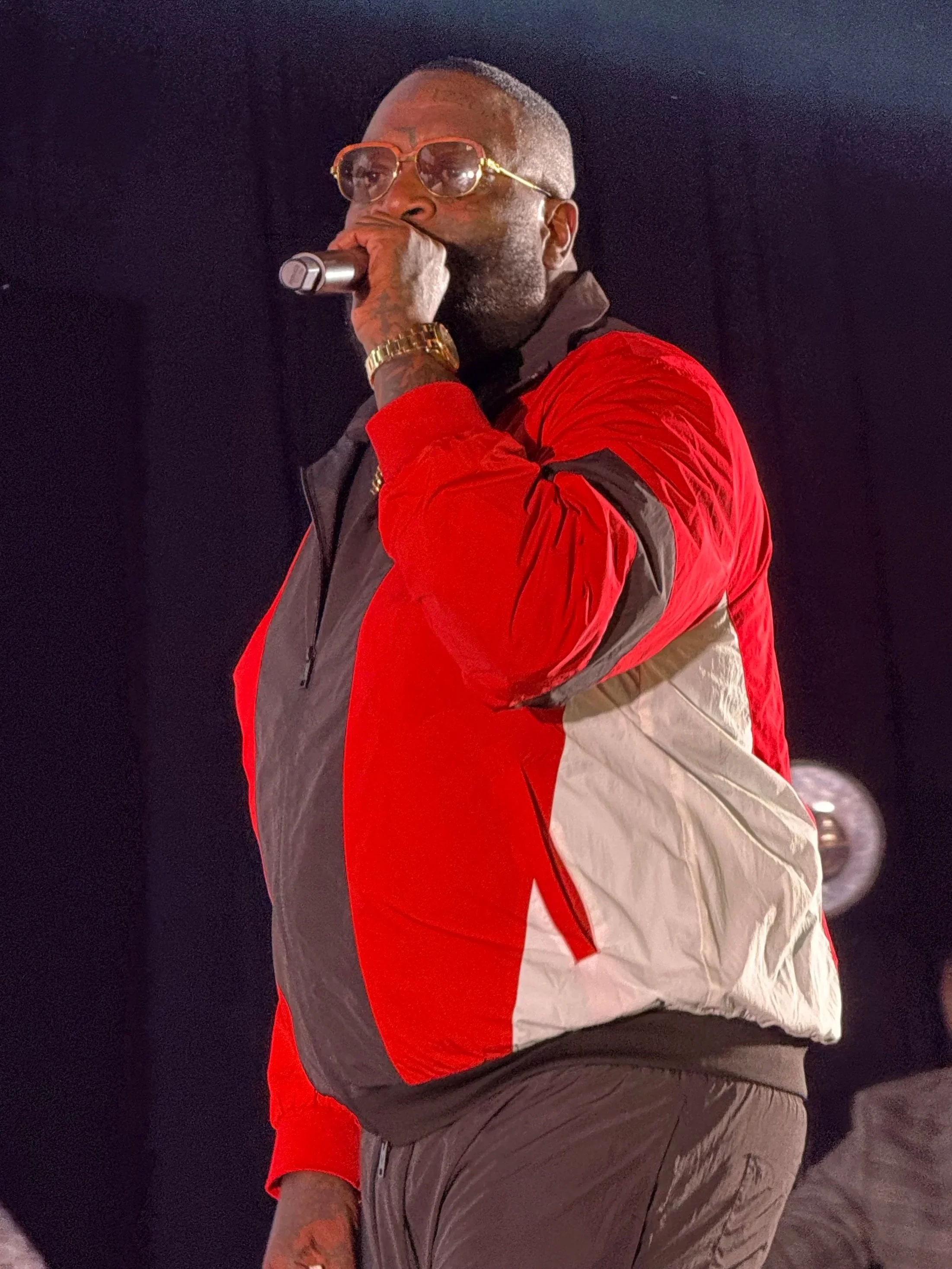 A man with gray hair, sunglasses, and a gold watch is holding a microphone and performing on stage. He is wearing a red, black, and beige jacket and brown pants.