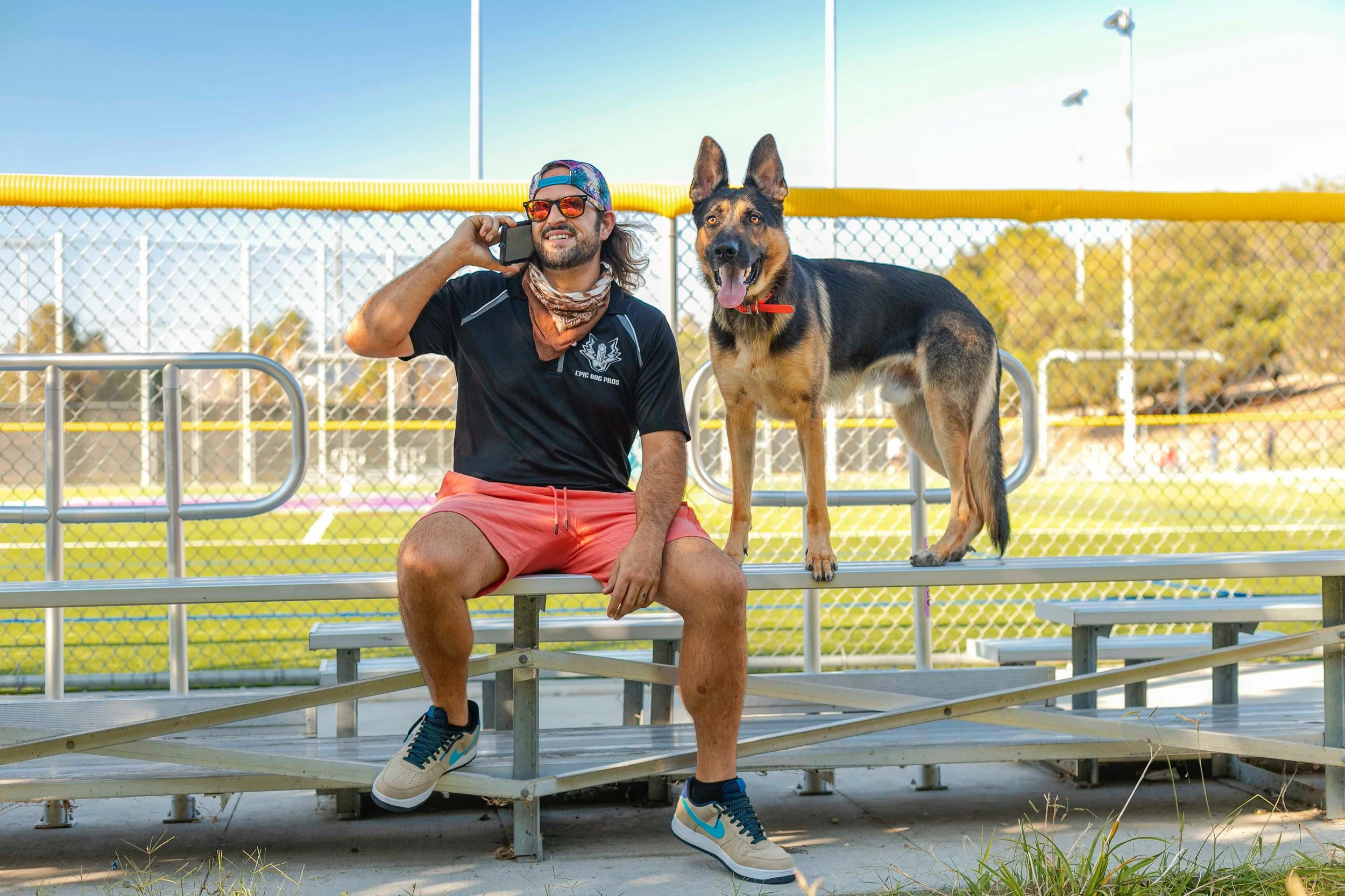 A man sitting on bleachers at a sports field, talking on a cell phone, with a large German Shepherd dog standing next to him.
