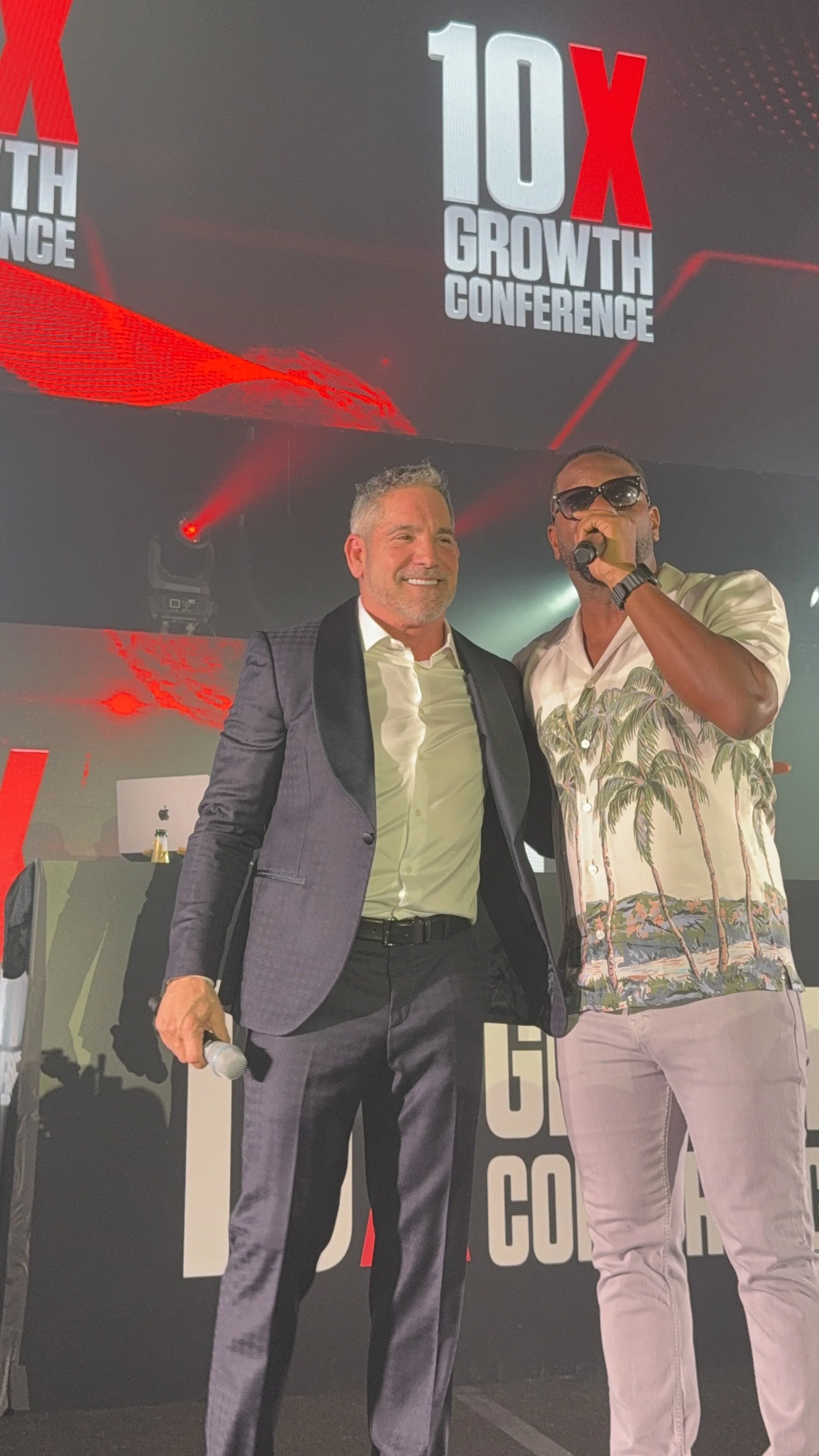 Two men standing on a stage under a large screen displaying the text '10X Growth Conference.' One man is smiling and dressed in a dark suit, white shirt, holding a microphone. The other man is wearing sunglasses, a Hawaiian shirt with palm trees, and
