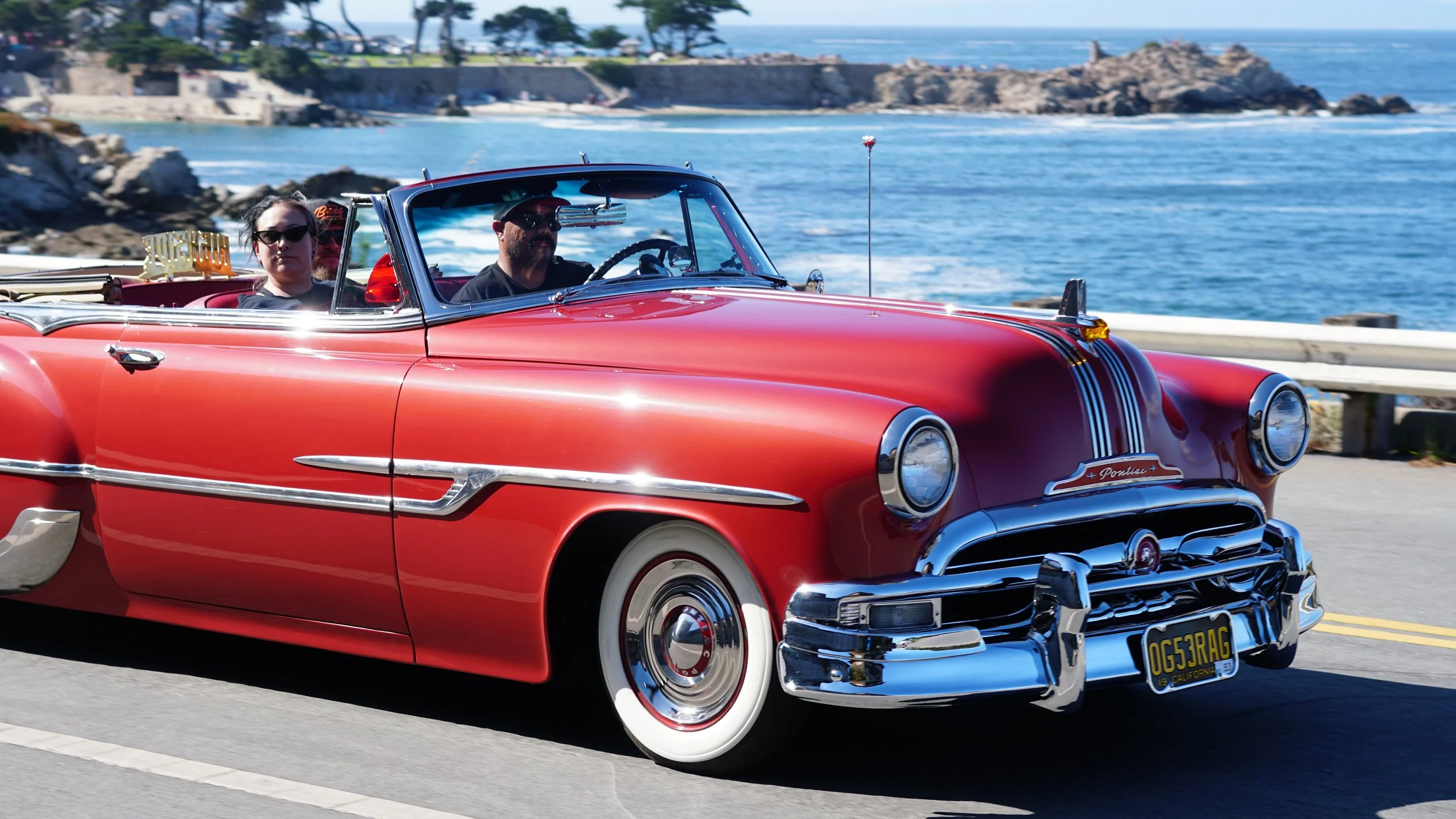A red vintage convertible car driving along the coast with three passengers, a woman and two men, near the ocean with rocks and buildings in the background.