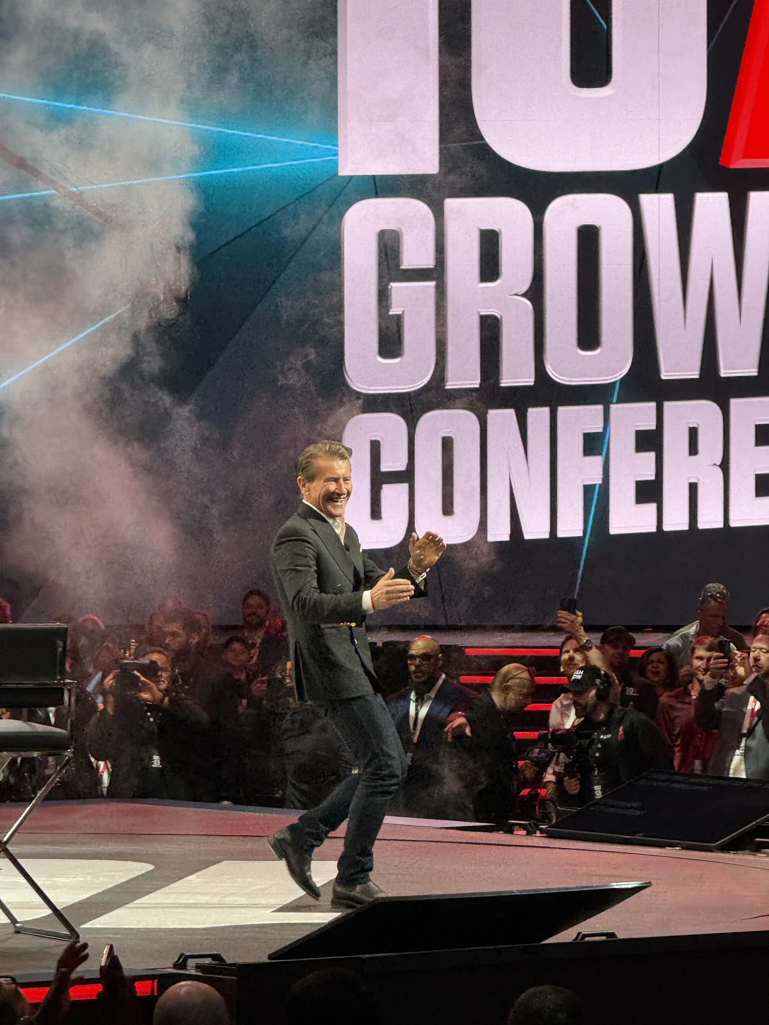A man in a black blazer and jeans on stage at a conference, smiling and clapping, with a large screen behind him displaying the words 'GROW CONFERENCE' and a crowd of people watching and taking photos.