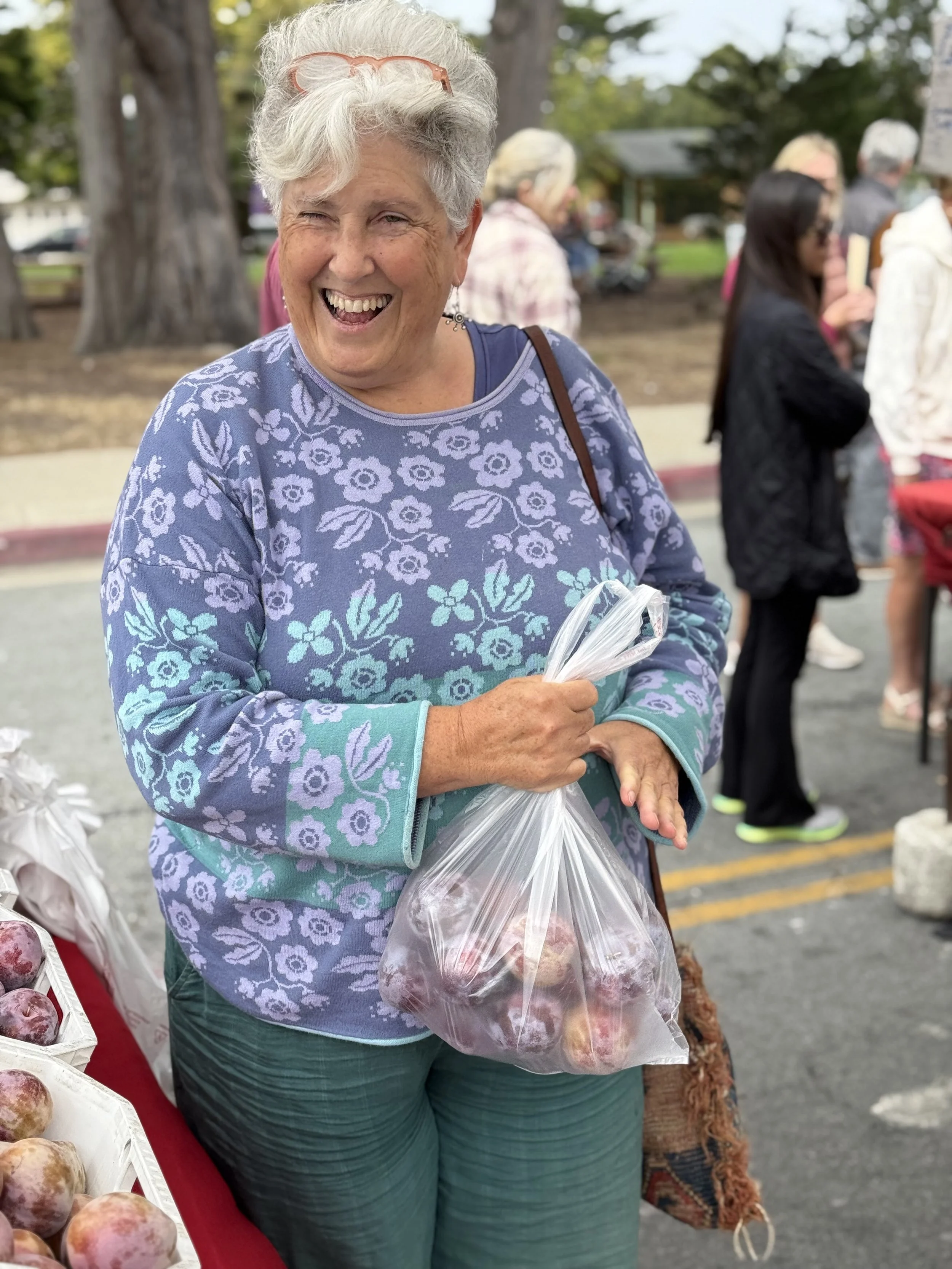 Smiling woman at an outdoor market holding a plastic bag of peaches.