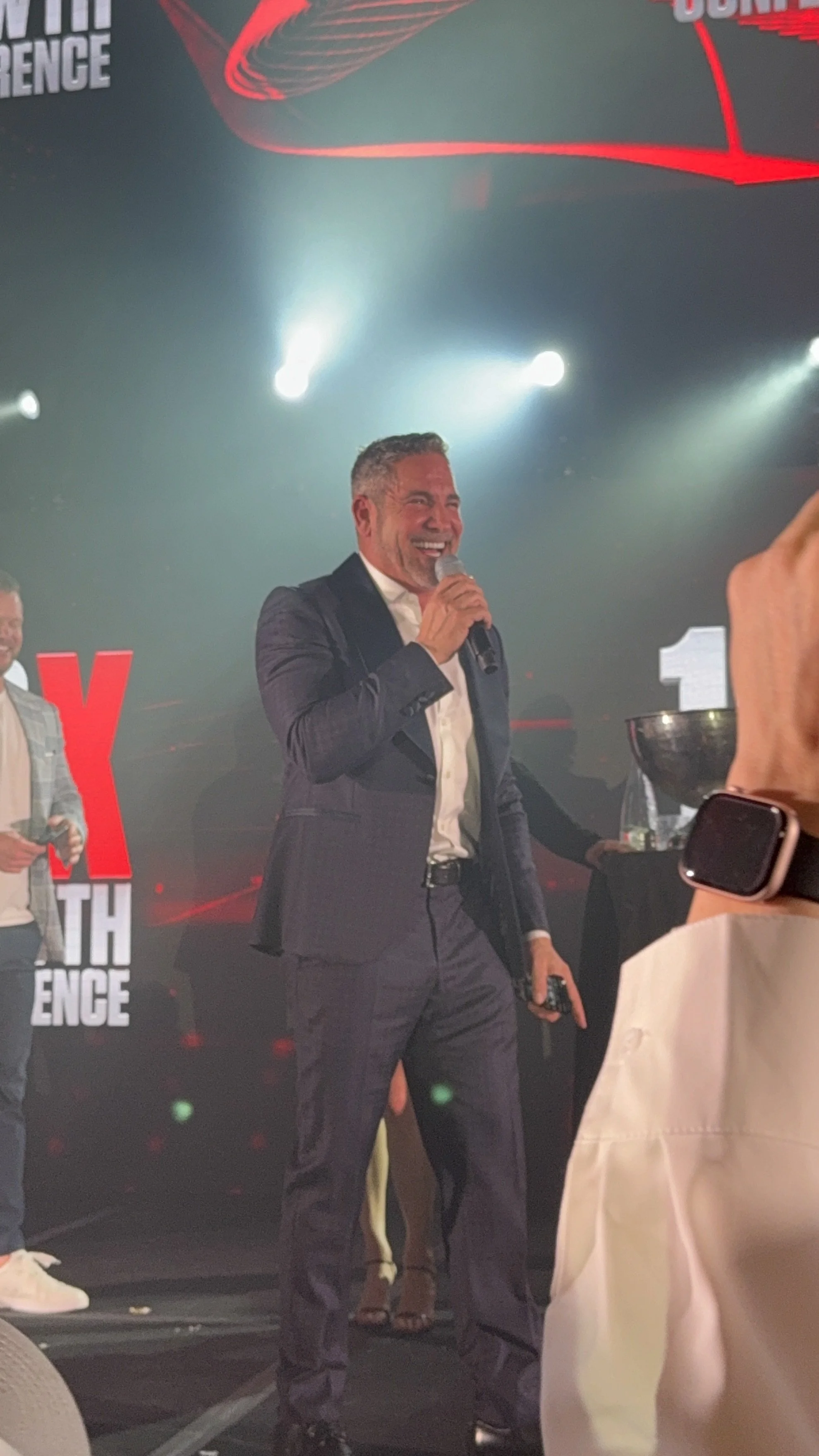 A man in a dark suit and white shirt is smiling and speaking into a microphone on stage at a professional event. There are stage lights and a large red and black backdrop with text that appears to say 'X' and 'Conference' behind him. Another man is v
