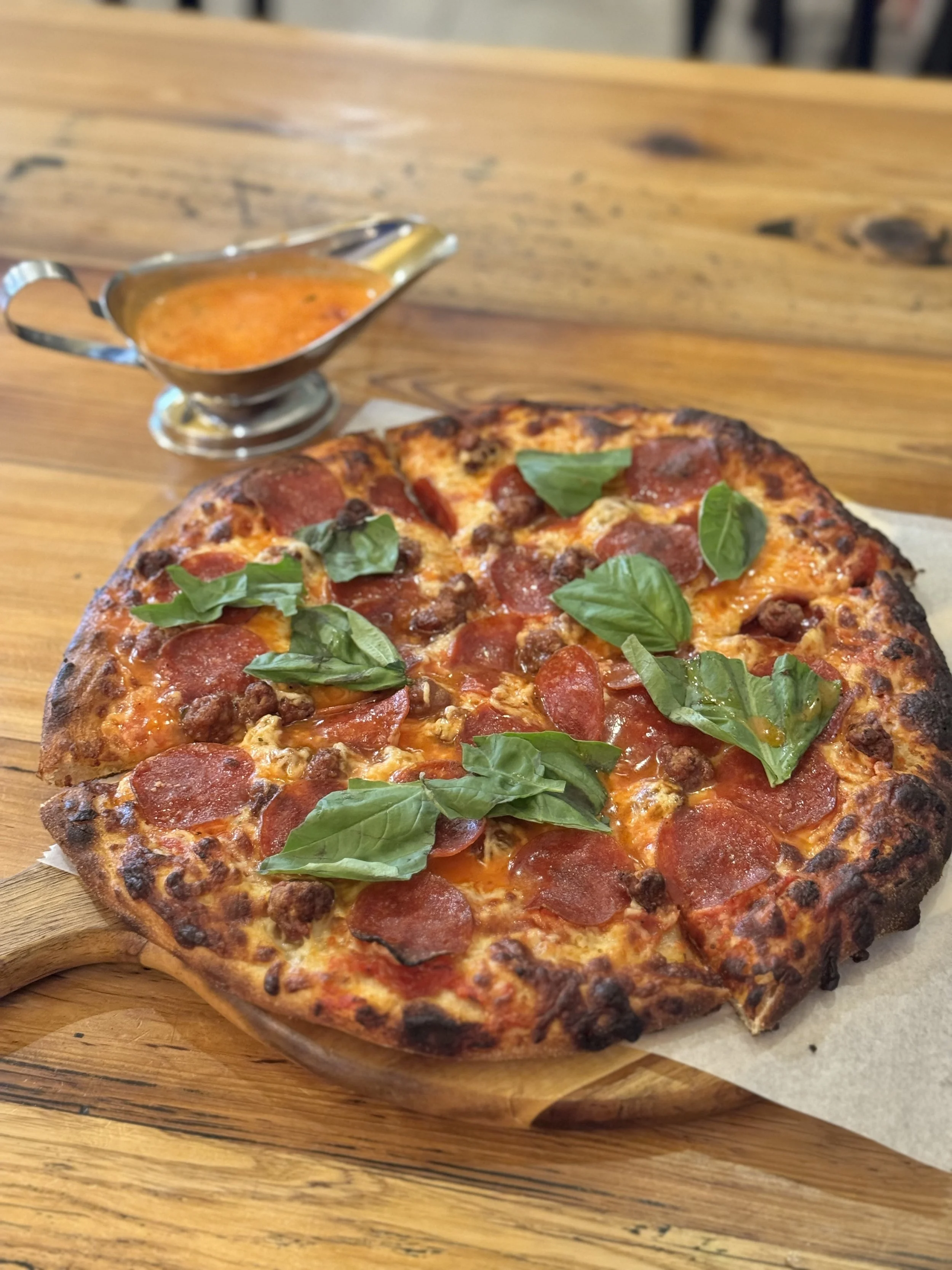 A pepperoni and sausage pizza topped with fresh basil leaves on a wooden serving board, with a side of marinara sauce in a silver gravy boat on a wooden table.