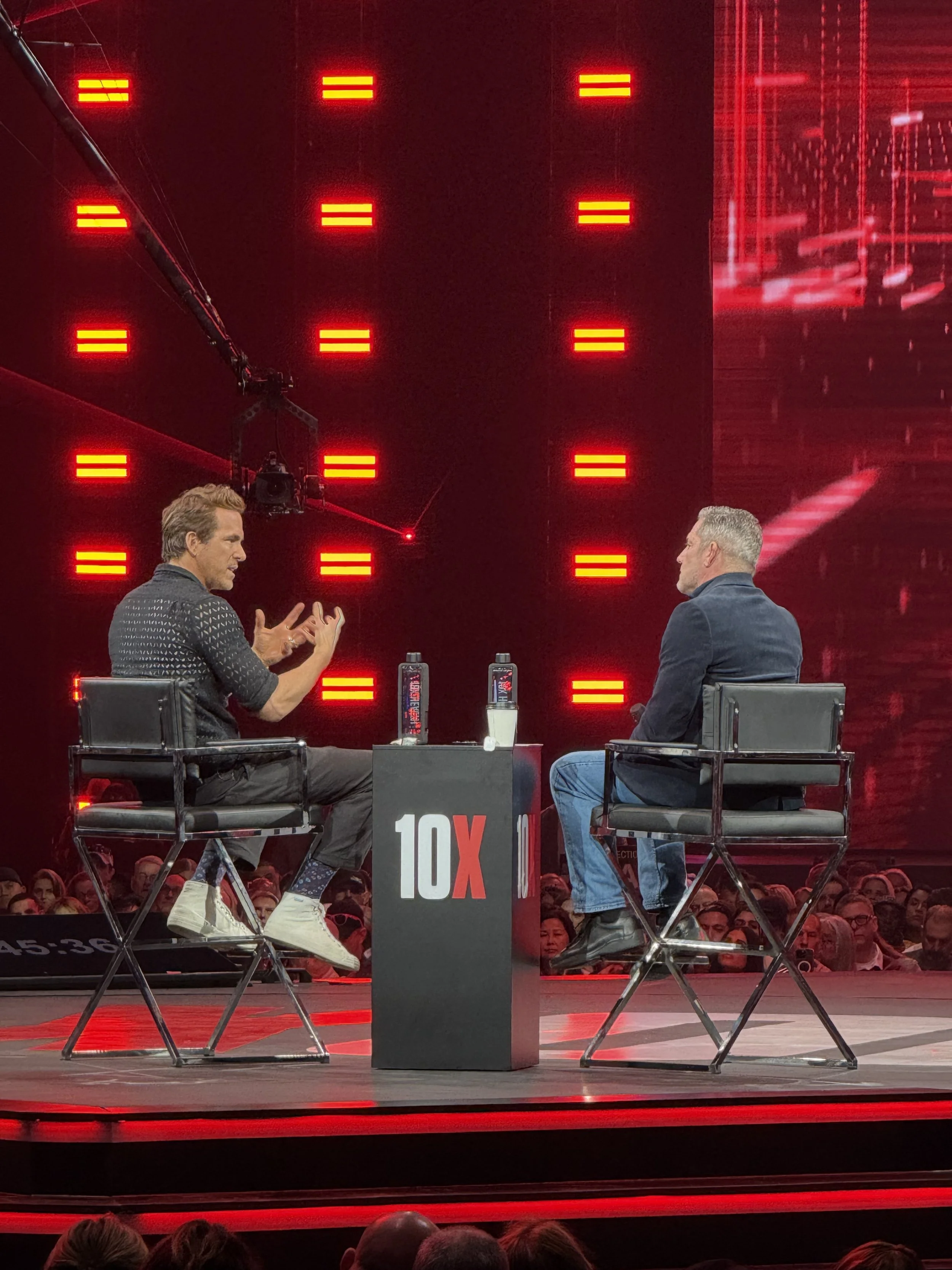 Two men sitting on stage in conversation during an event, with a red-lit backdrop, a black table with 10X on it, and audience members visible in the background.
