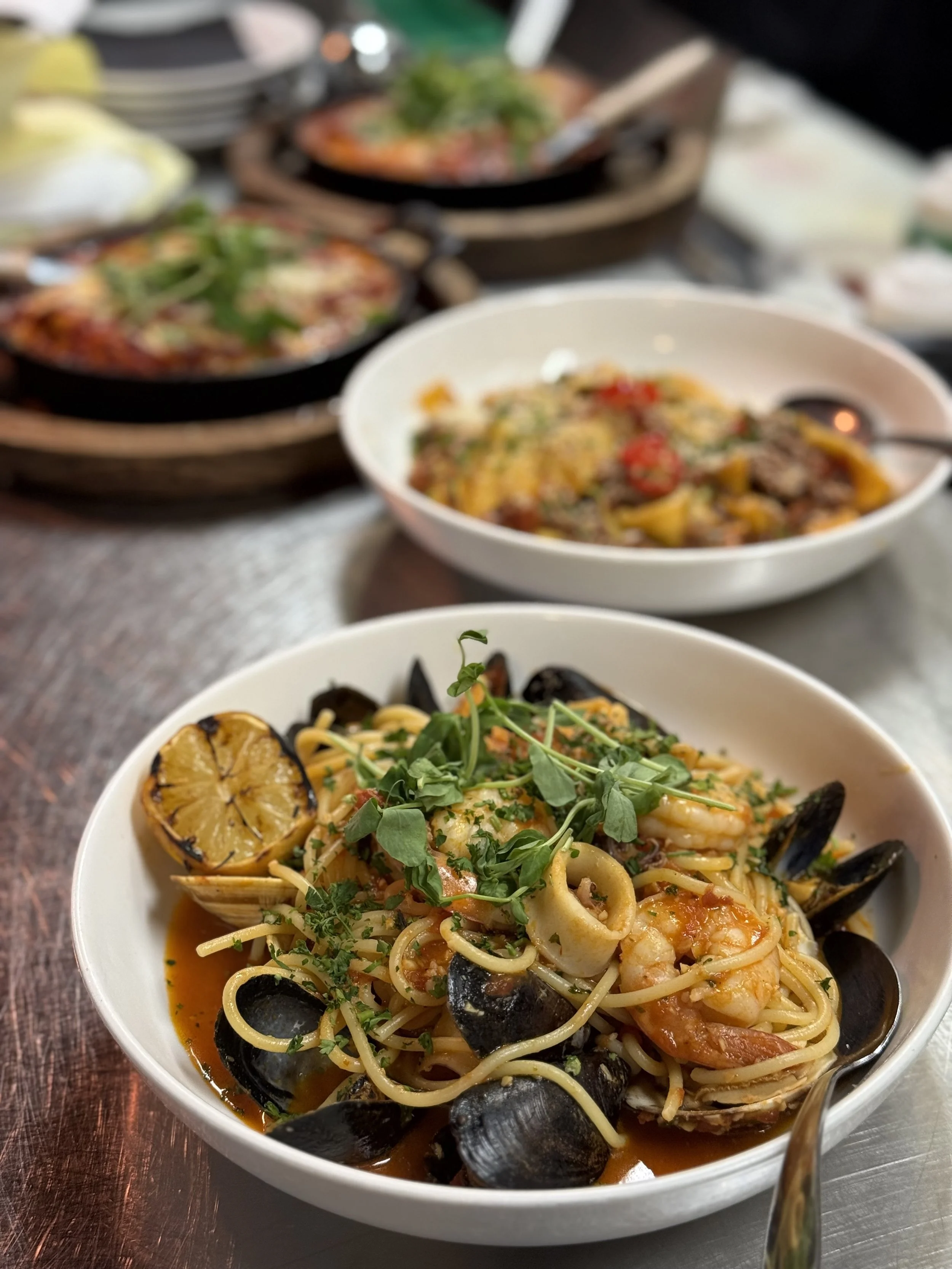 A bowl of seafood pasta garnished with fresh herbs, with lemon halves and mussels. In the background, there are two other dishes, possibly pizza and another pasta, on a wooden table.