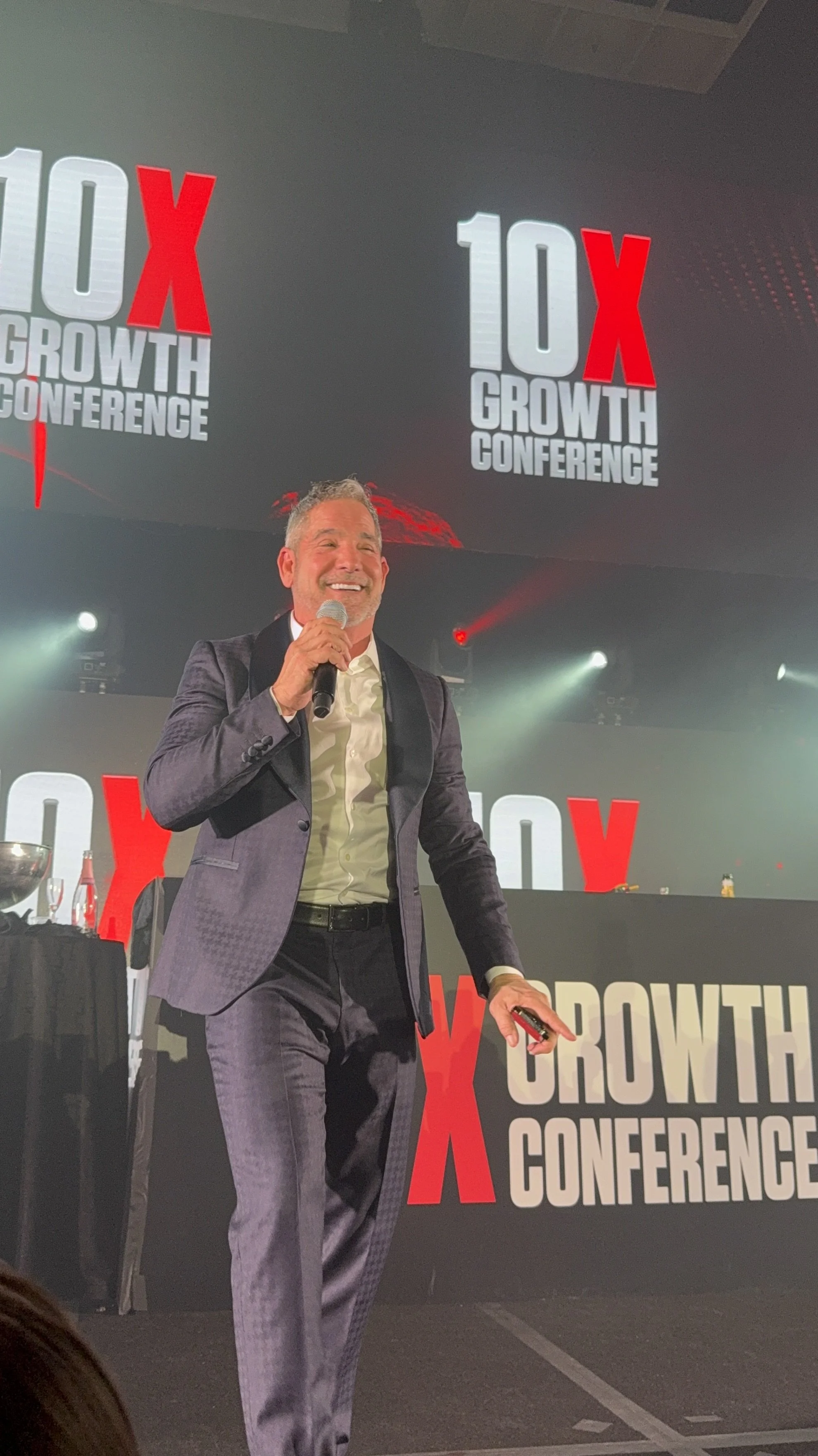 A man in a suit smiling and holding a microphone on stage at the 10X Growth Conference with large screens displaying the event logo in the background.