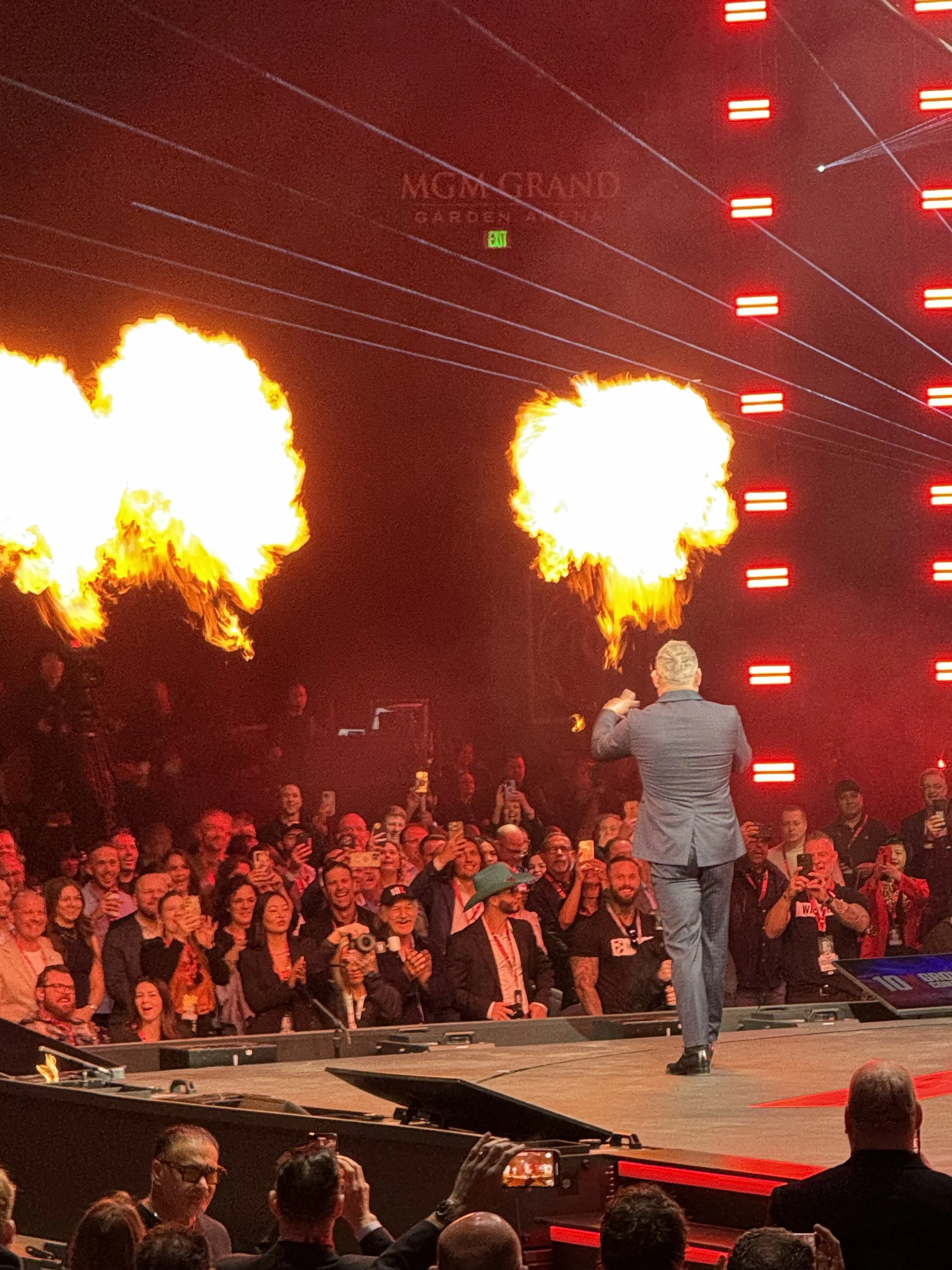 A man in a suit performing a fire-breathing act on stage during a presentation or show at MGM Grand Garden Arena with a crowd watching and taking photos.