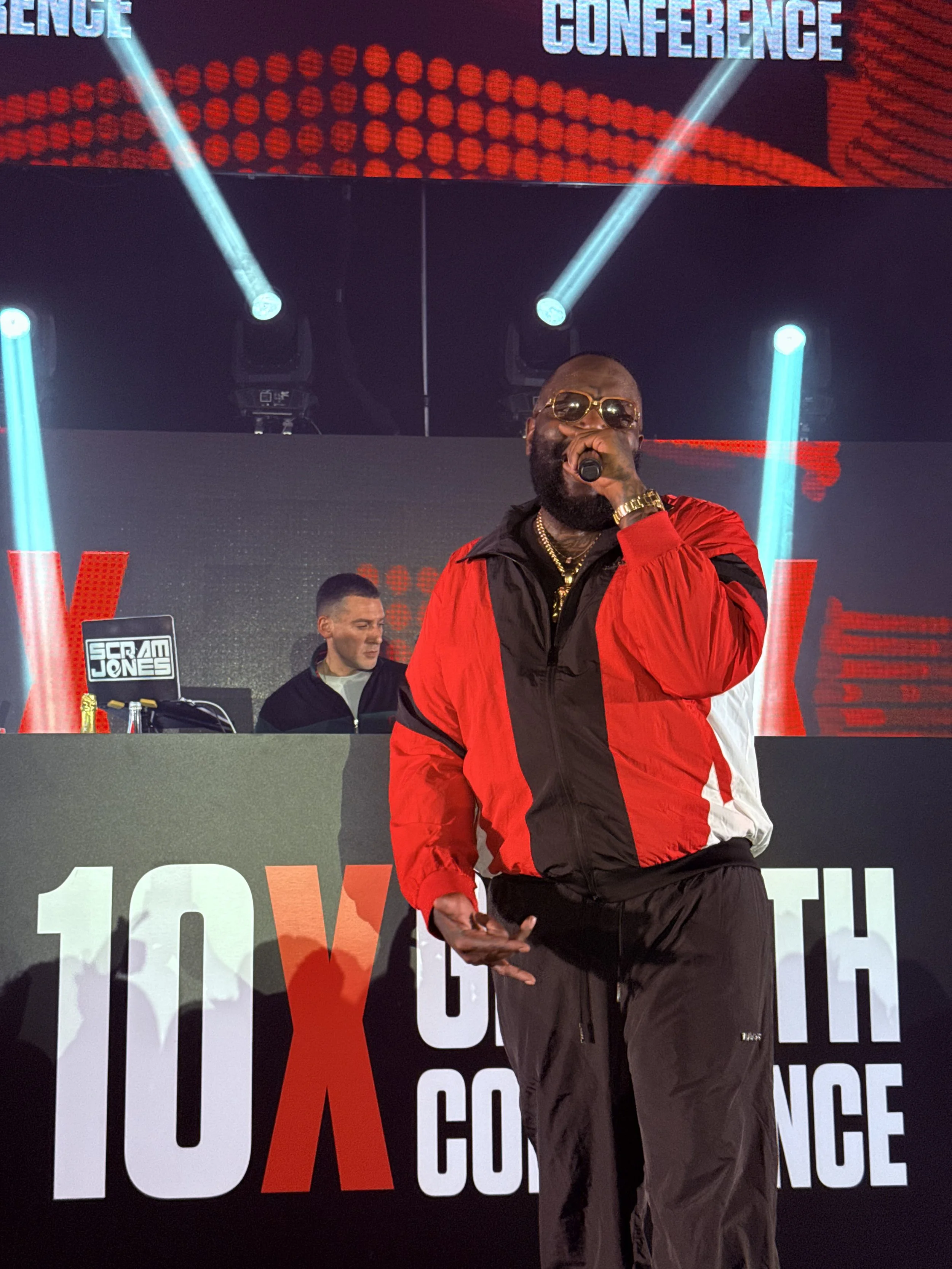 Music performer in a red and black tracksuit with gold jewelry and sunglasses singing into a microphone at a 10X conference stage, with a DJ behind him and stage lights.
