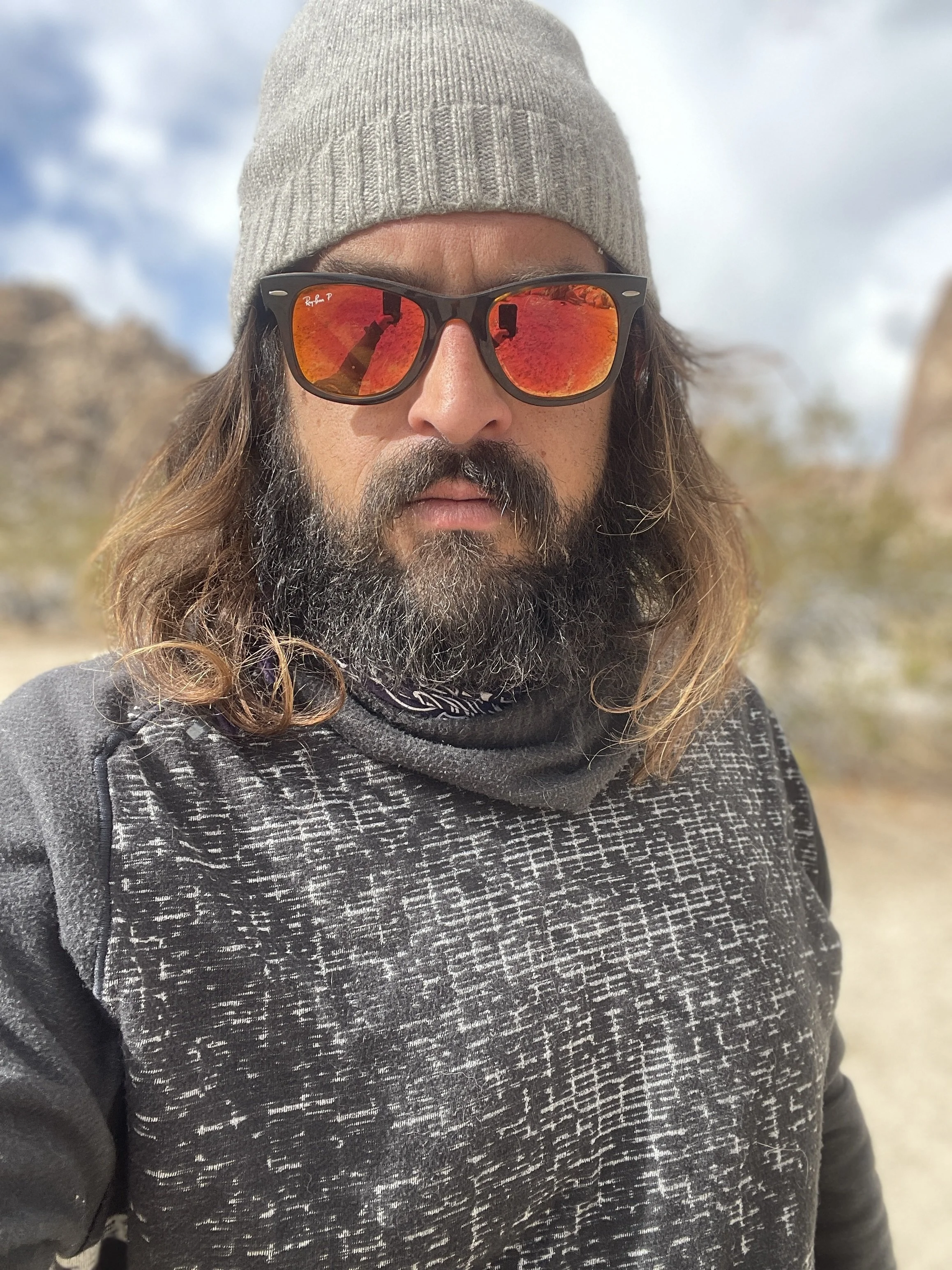 Man with long hair and beard wearing a gray beanie, sunglasses with orange mirrored lenses, a patterned gray shirt, and a neck gaiter, outdoors in a desert-like environment.
