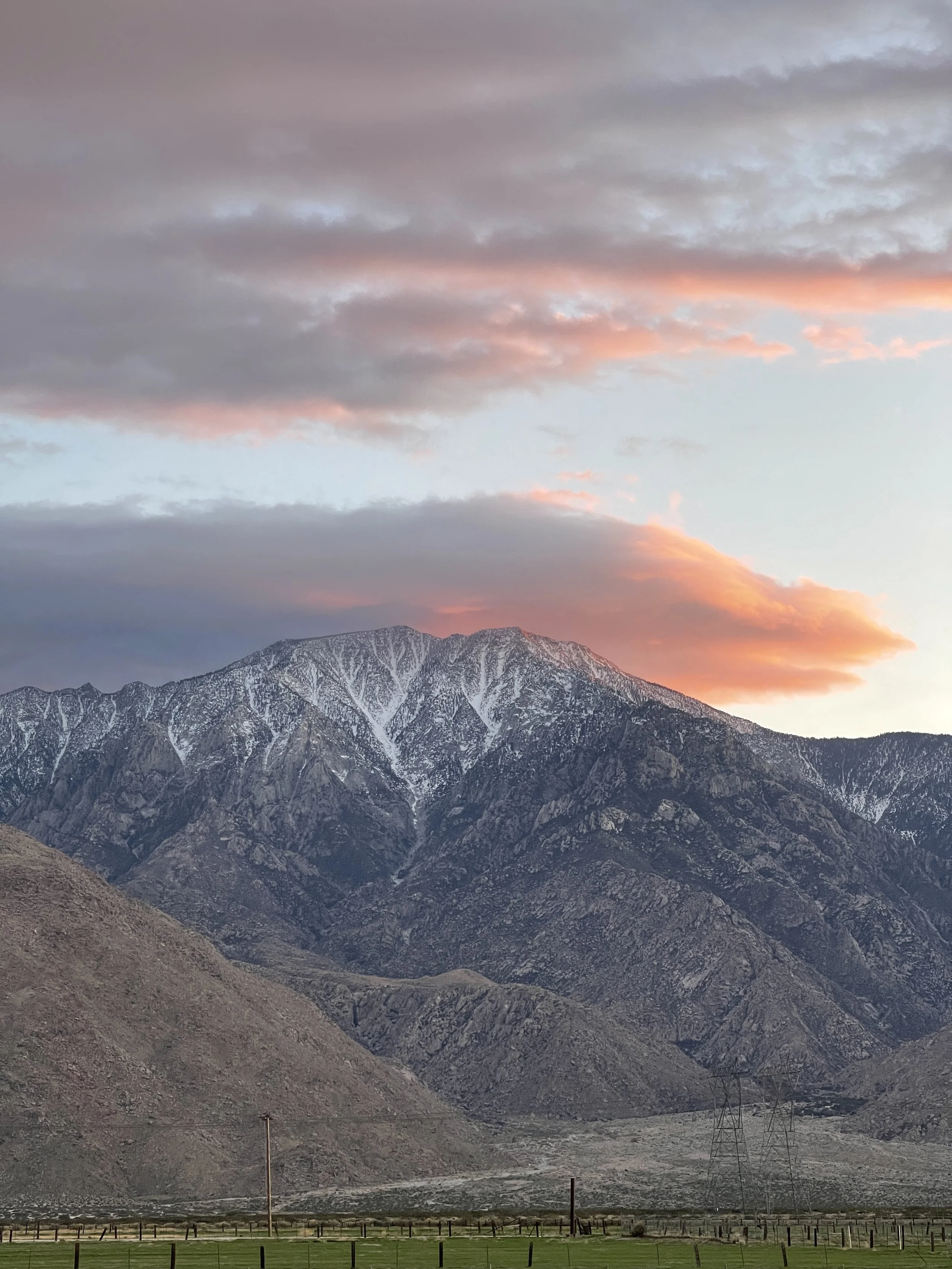 Mountain range with snow-capped peaks under a colorful sunset sky, with power lines in the foreground.