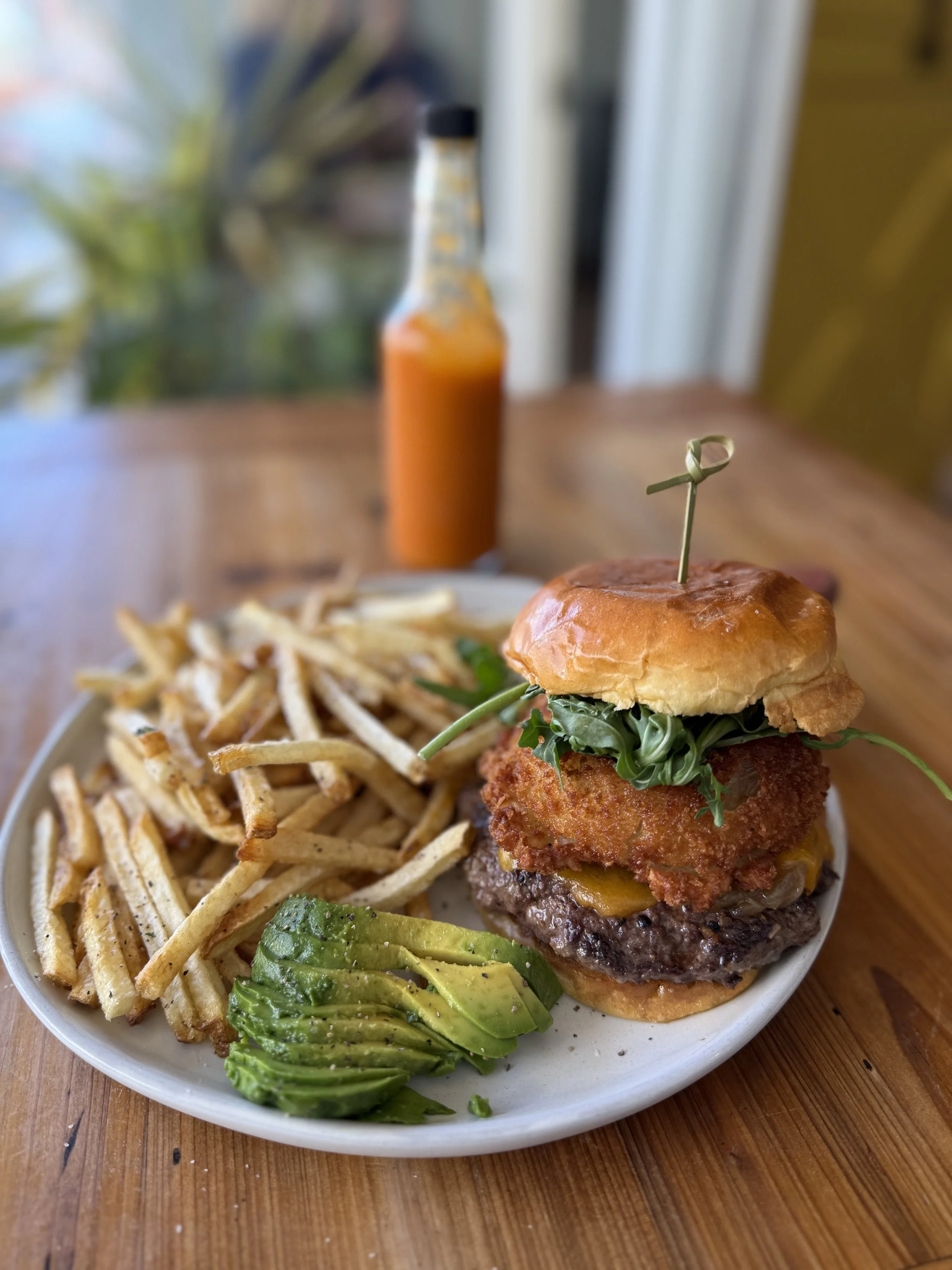 A plate with a cheeseburger topped with a fried chicken fillet, leafy greens, and a bun, served with French fries and sliced avocado, and a bottle of orange sauce in the background on a wooden table.