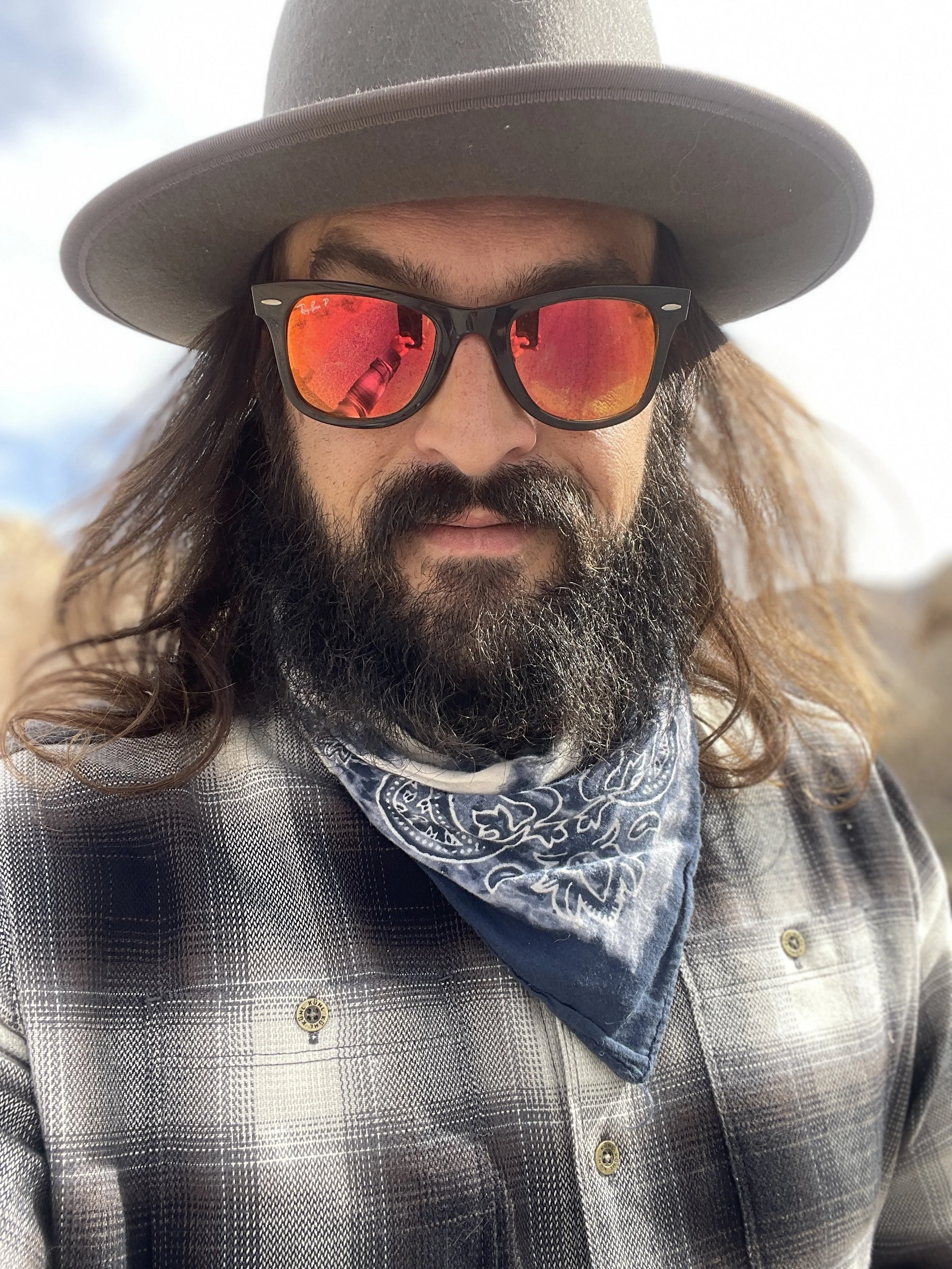 A man with long hair and a beard wearing a wide-brimmed hat, sunglasses with red reflective lenses, a blue bandana around his neck, and a plaid shirt outdoors.