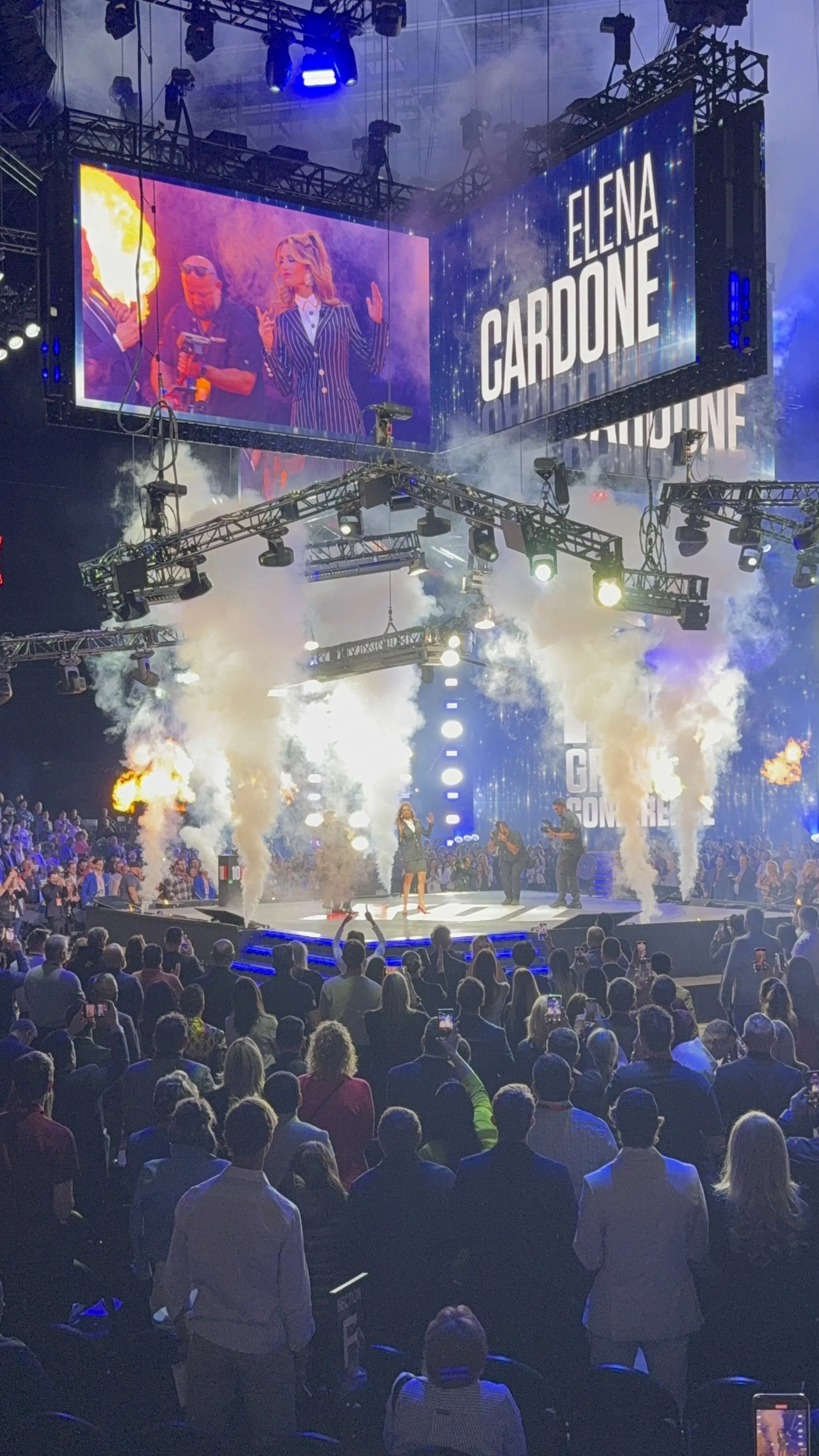 Elena Cardone speaking on stage at a conference with pyrotechnics and smoke effects, large screens displaying her name, and a crowd of attendees watching.