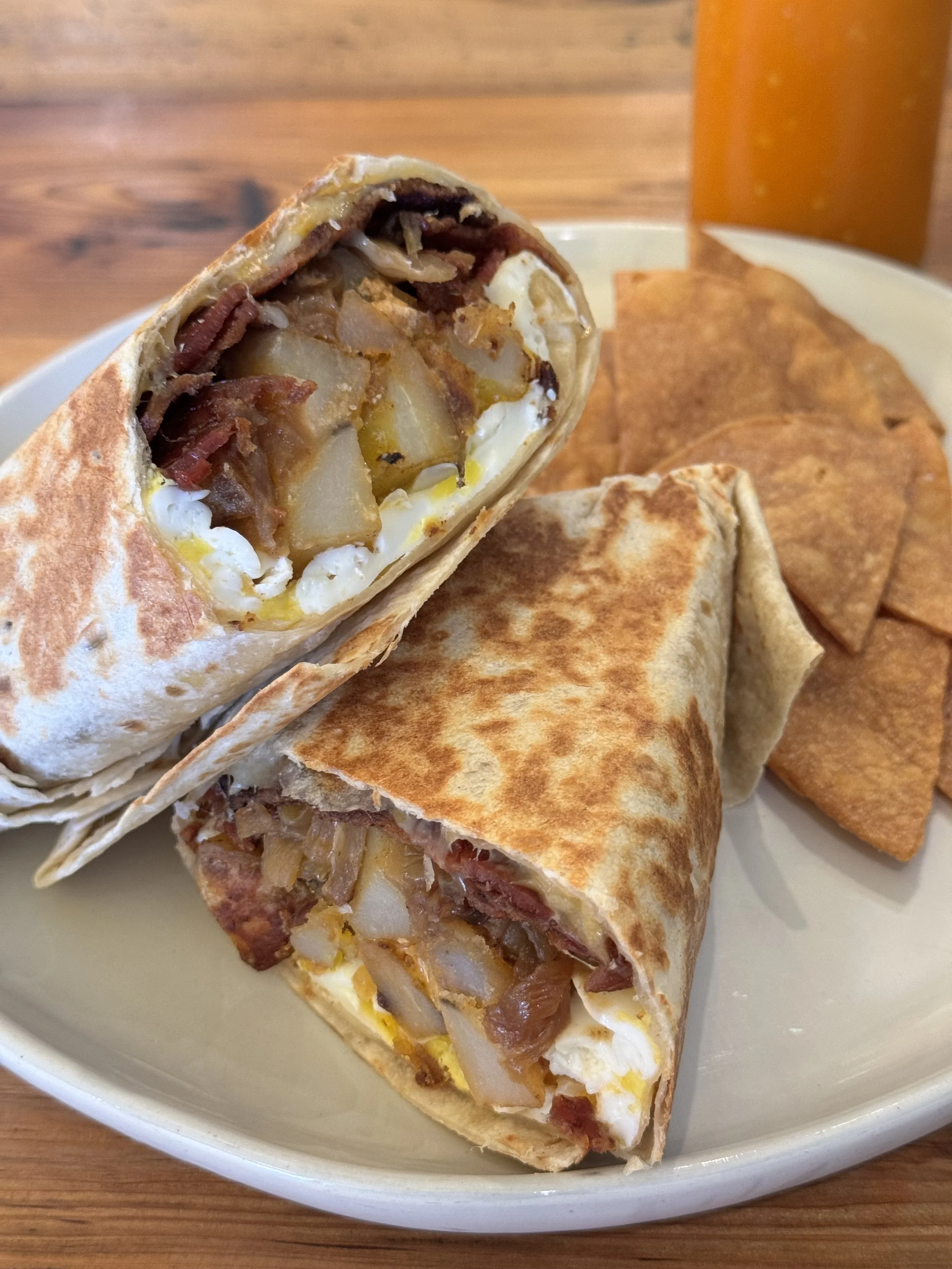 A plate with a breakfast burrito cut in half showing potatoes, eggs, bacon, and cheese inside, accompanied by tortilla chips and a drink in the background.