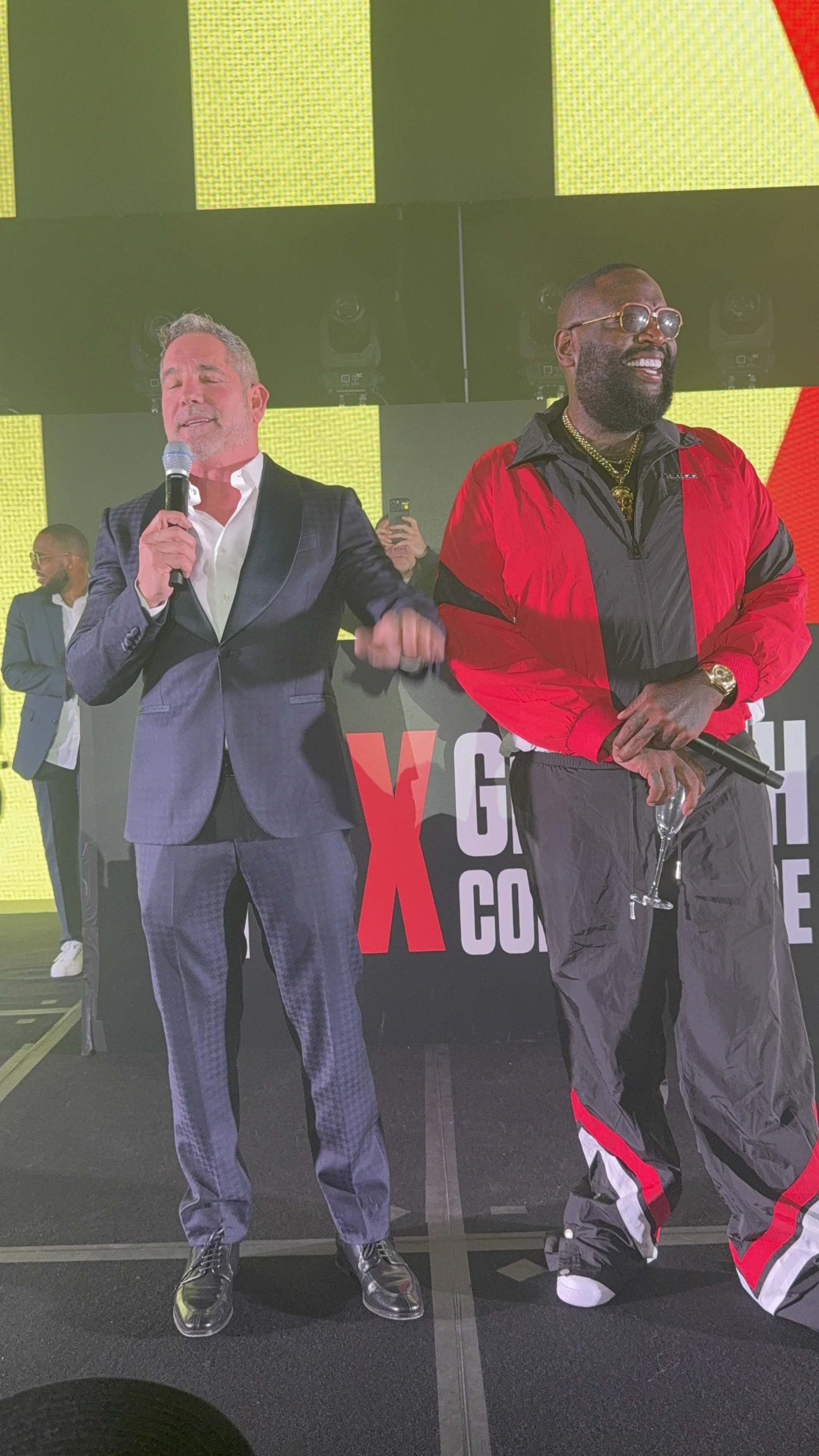 Two men standing on stage at a TEDx event, holding microphones. The man on the left is wearing a dark suit and white shirt, and the man on the right is dressed in a red and black athletic tracksuit with sunglasses and gold jewelry.