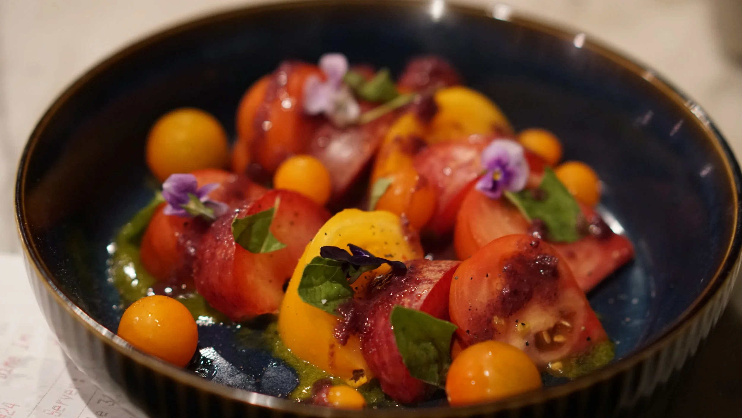 A black bowl contains a colorful salad with cherry tomatoes, sliced peaches, small yellow tomatoes, green basil leaves, and edible purple flowers, with a drizzle of dressing.