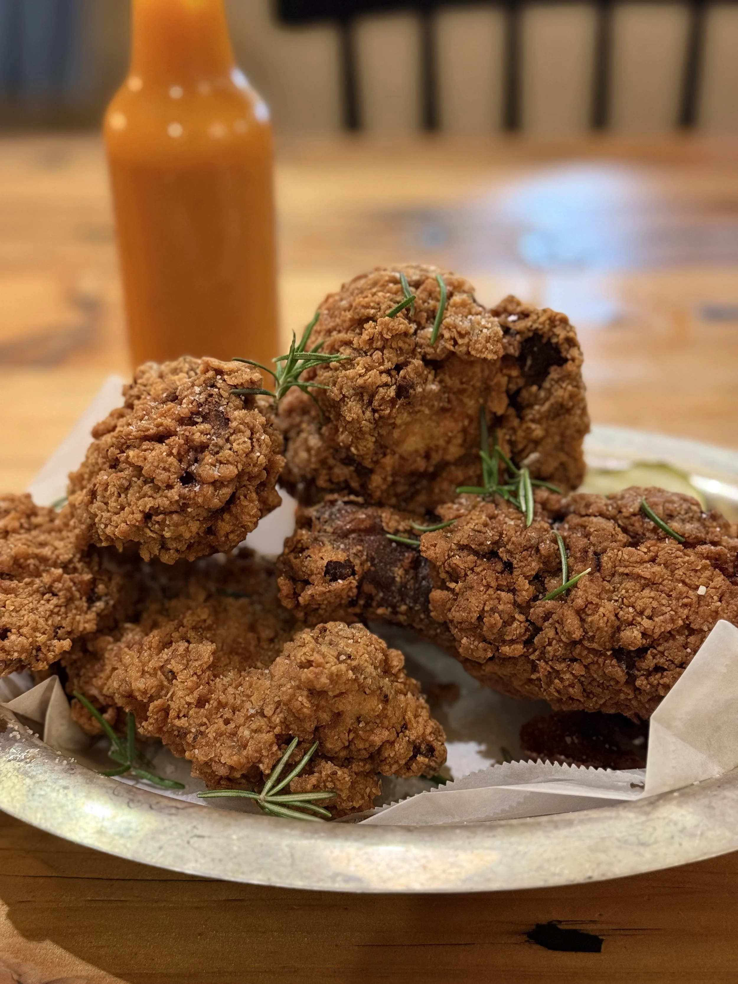 Plate of fried chicken pieces garnished with fresh herbs, with a bottle of hot sauce in the background on a wooden table.
