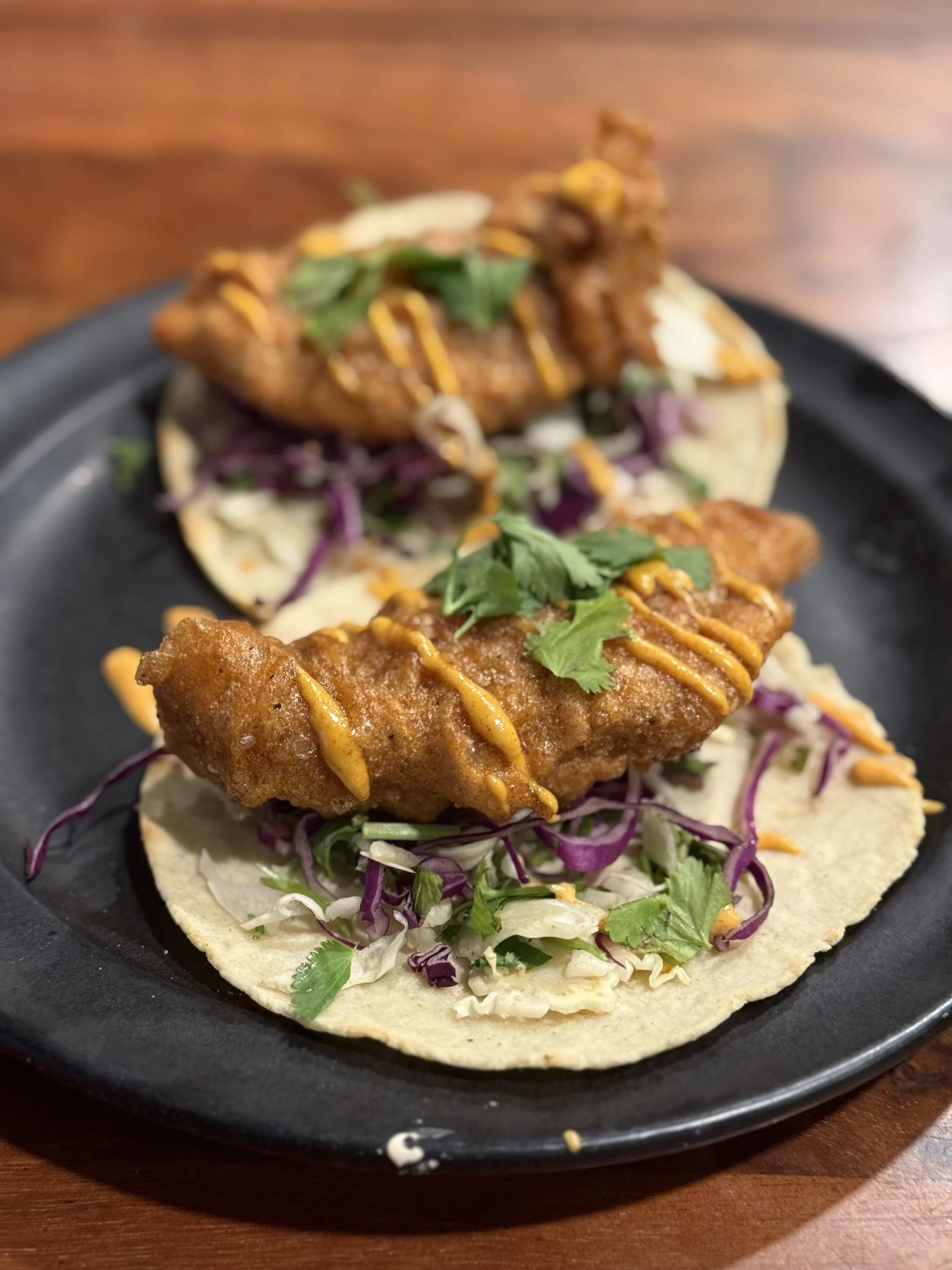 Two fish tacos on a black plate, topped with cilantro, drizzled with orange sauce, with shredded cabbage and purple cabbage on soft corn tortillas.