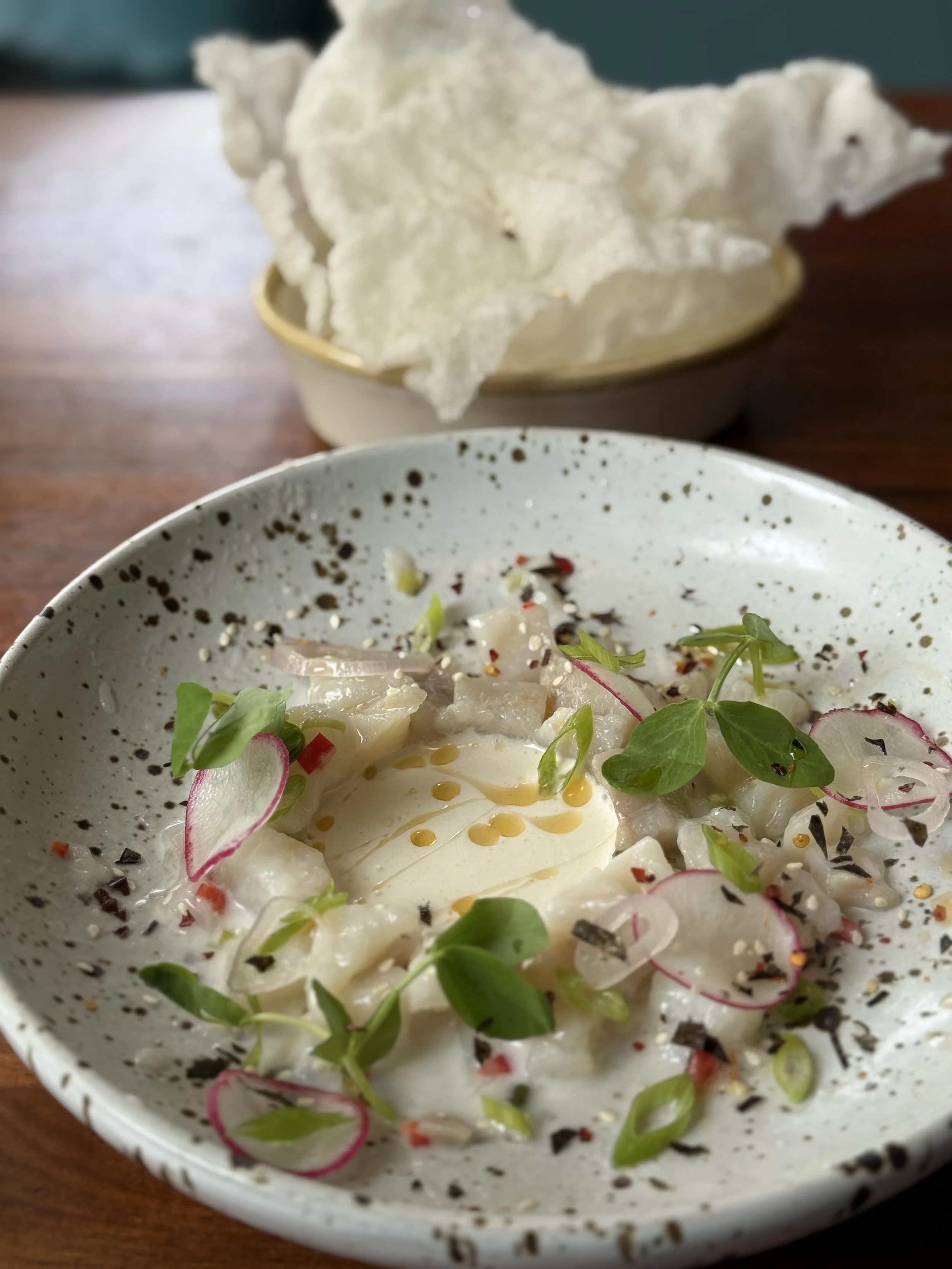 A bowl with sliced raw fish, herbs, and spices, topped with a soft-boiled egg, with a bowl of prawn crackers in the background.