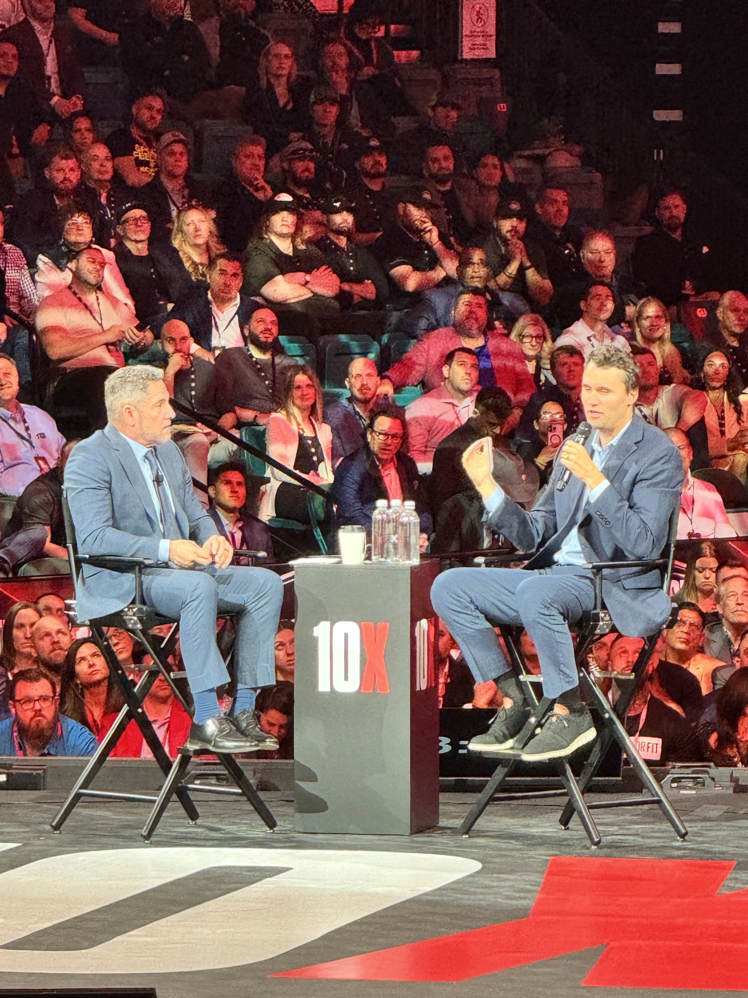 Two men are seated on stage in conversation during a panel event for TEDx. They are dressed in suits and holding microphones. The stage has a black table with two water bottles and a cup, and the audience is visible in the background under red stage