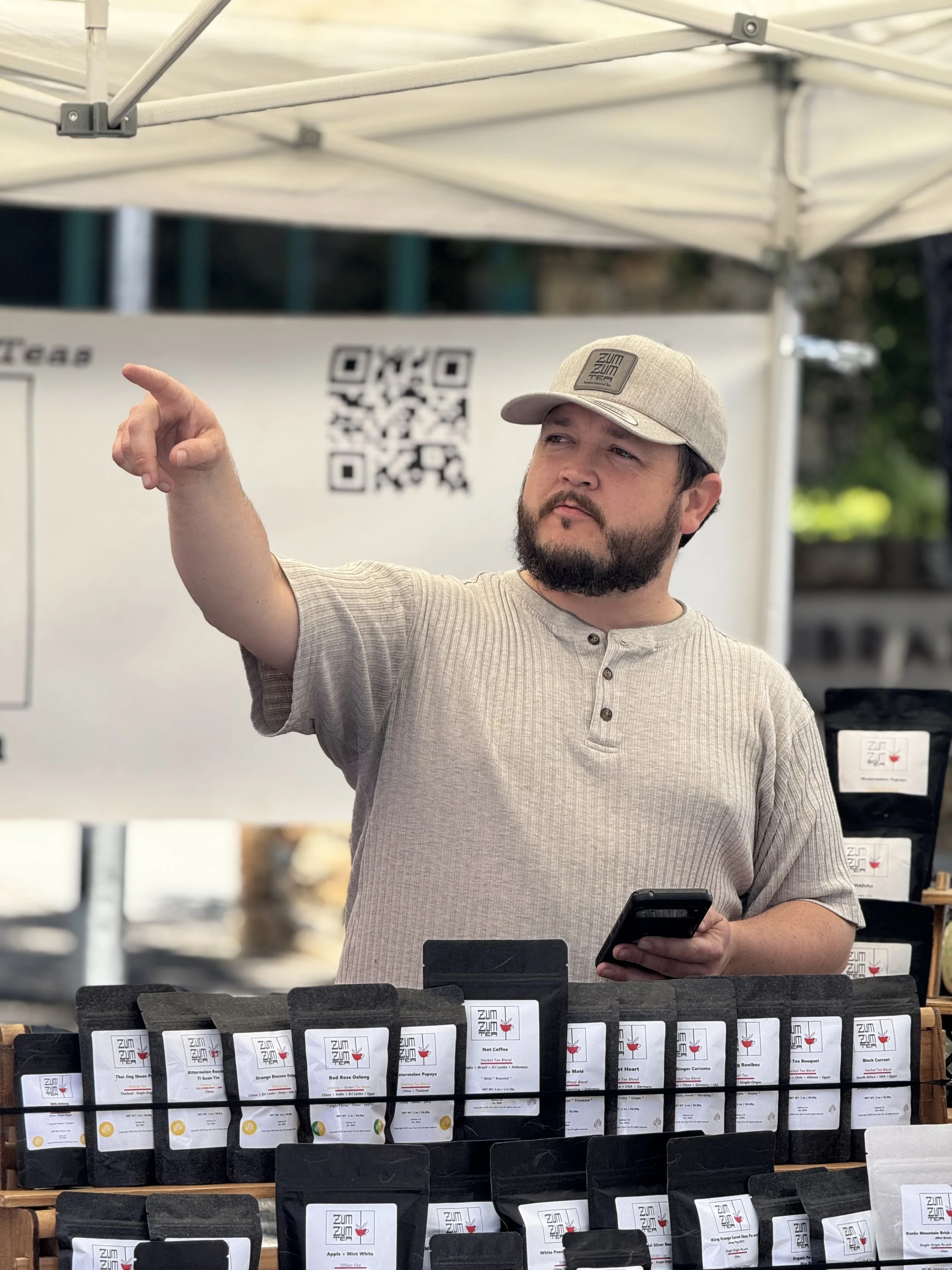 A man at an outdoor market stand, wearing a beige cap and a beige short-sleeved shirt, is pointing to his left while holding a smartphone in his right hand. The stand has several black packages of products with white labels, and there is a QR code on