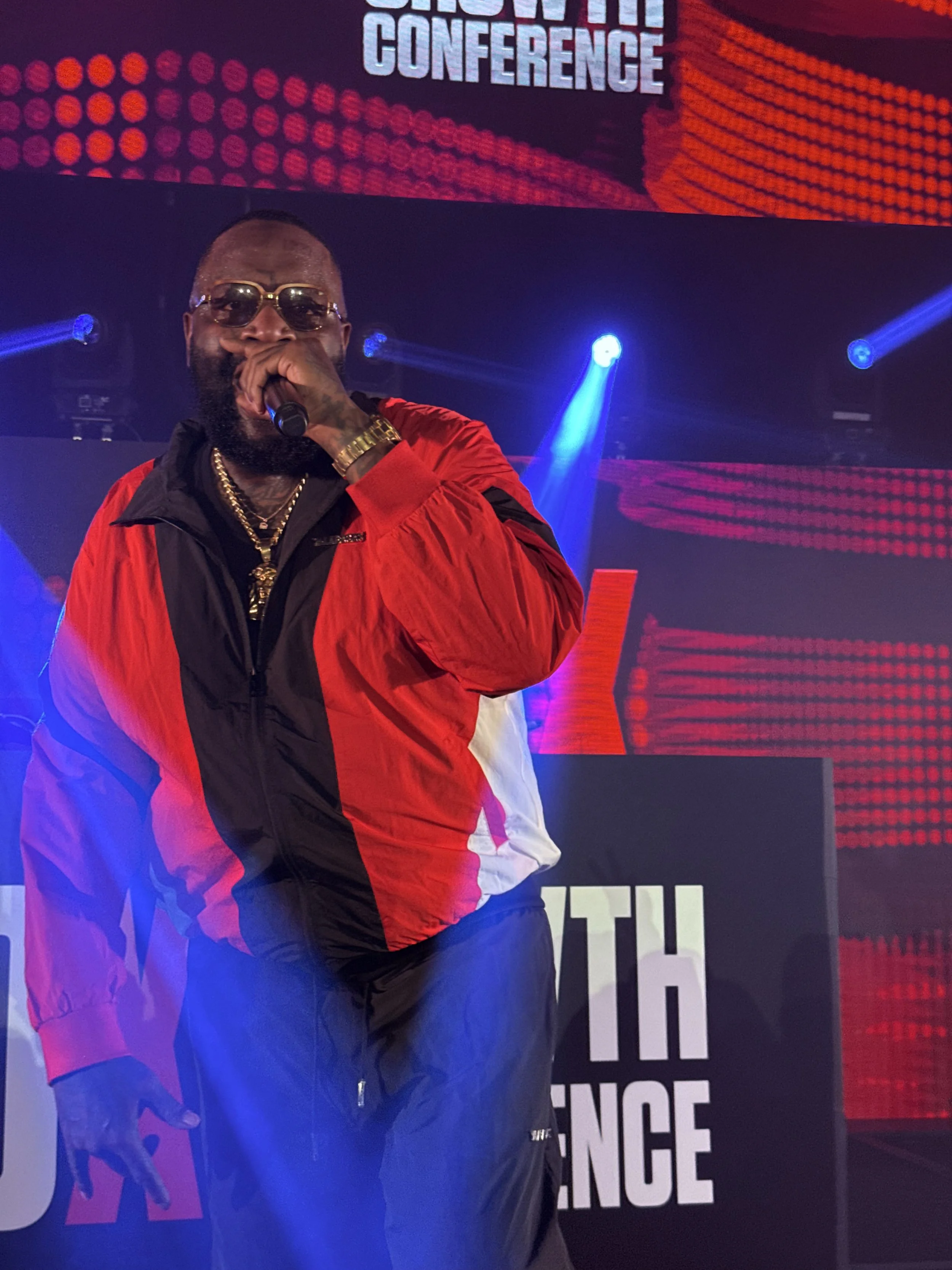 A man wearing sunglasses, a red and black jacket, gold jewelry, and a gold watch is holding a microphone on stage at a music event with bright blue and purple lighting effects and a large screen displaying the words 'SHOW THE CONFERENCE' in the backg