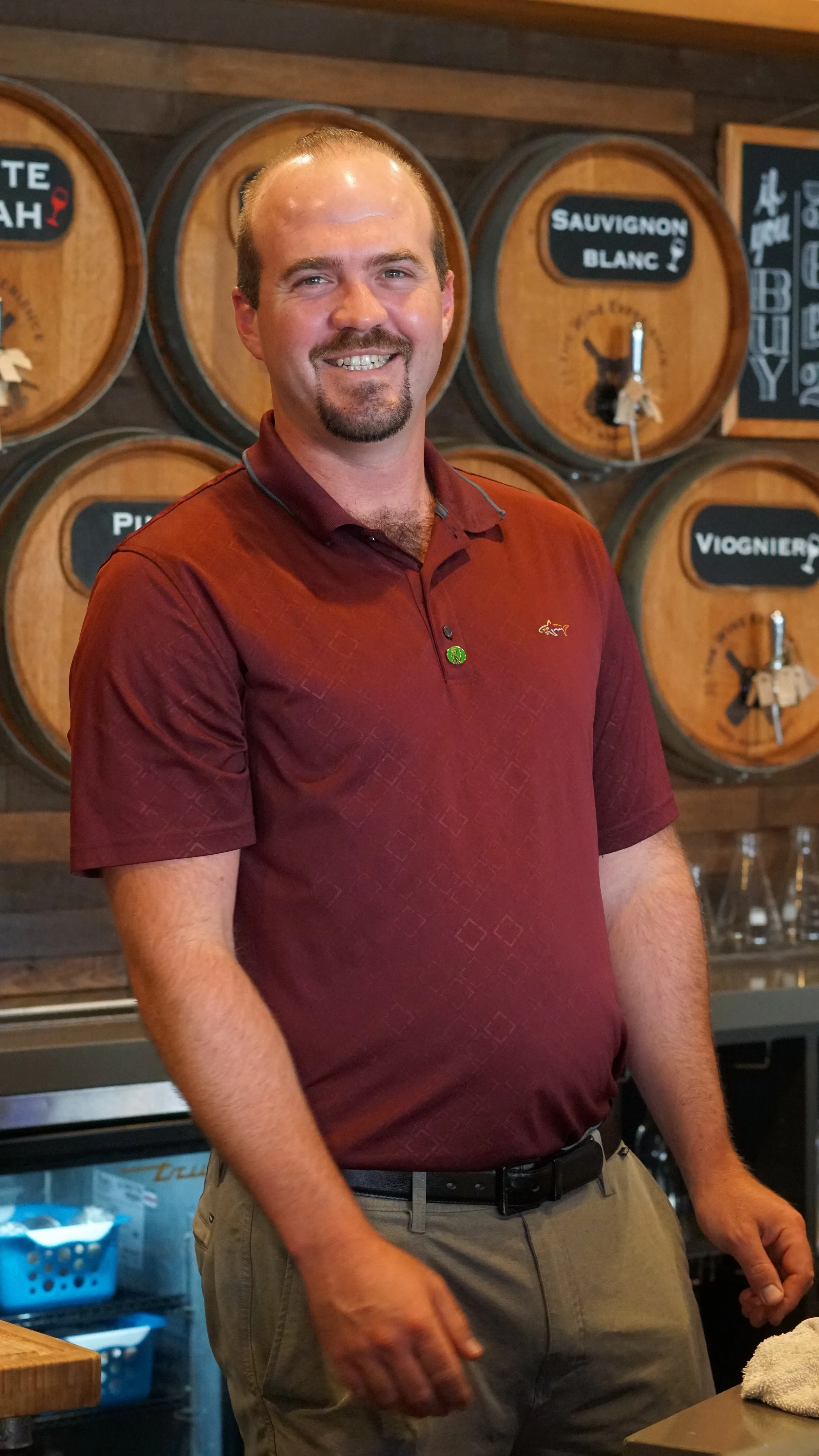 A man with light skin, short hair, and a goatee smiling, wearing a maroon polo shirt and khaki pants, standing indoors in front of wooden wine barrels with labels, in a wine bar or restaurant setting.