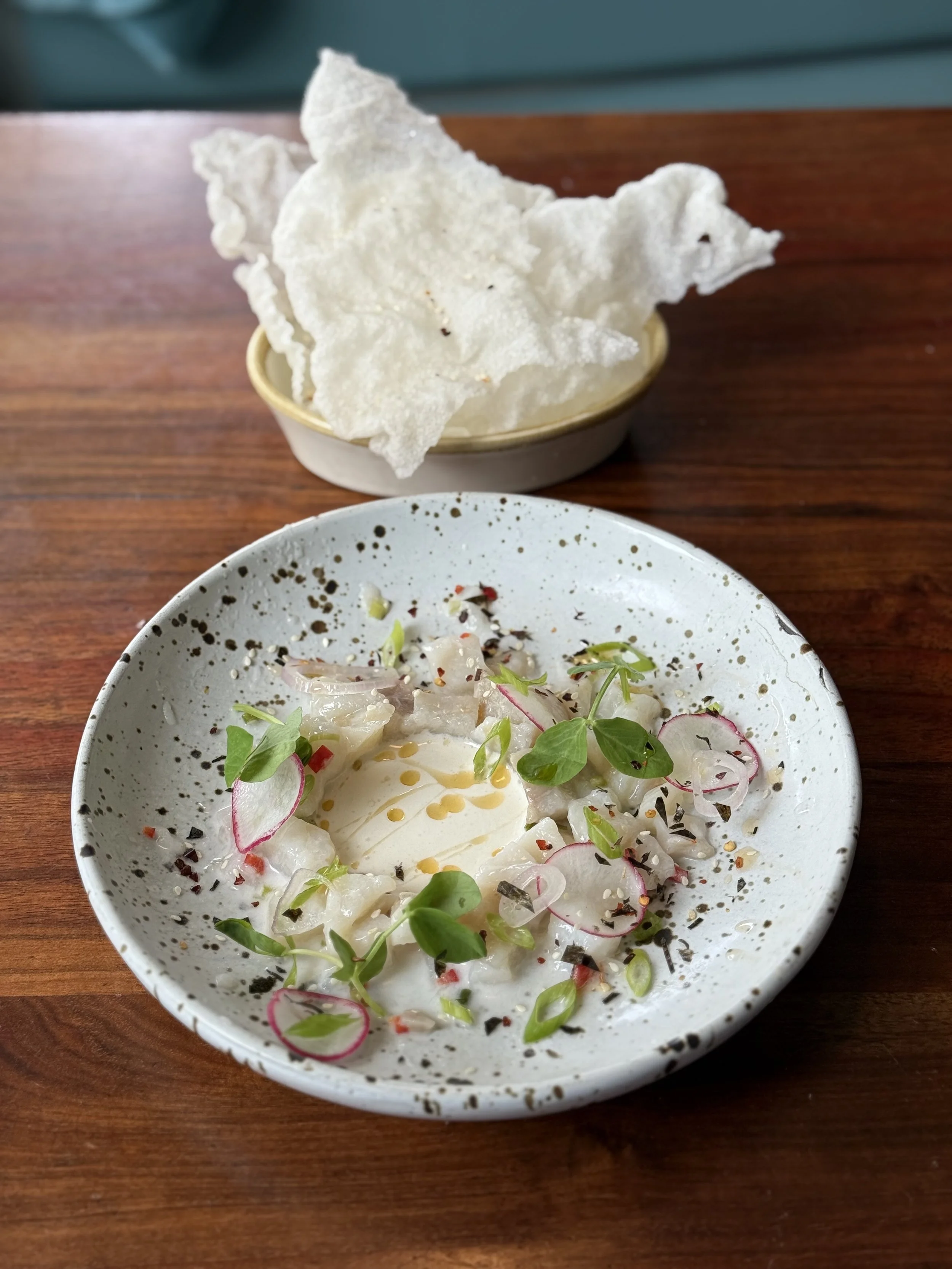 Ceviche served in a white bowl garnished with radish slices, microgreens, and a drizzle of oil, with crispy tortilla chips in a separate bowl behind.