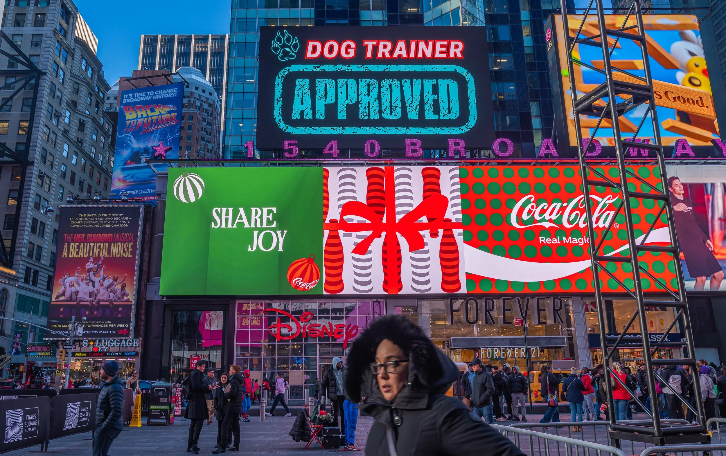 Timely Times Square billboard displays various advertisements. The top features a digital sign with the word "APPROVED" in blue, with changeable text above and below including "DOG TRAINER." Below is a large Coca-Cola ad with festive Christmas theme, a gift box, and the slogan "Share Joy." The Disney logo is visible on the building's façade, and there are numerous people walking or standing around, some bundled in winter clothing, on a busy city street during evening hours.