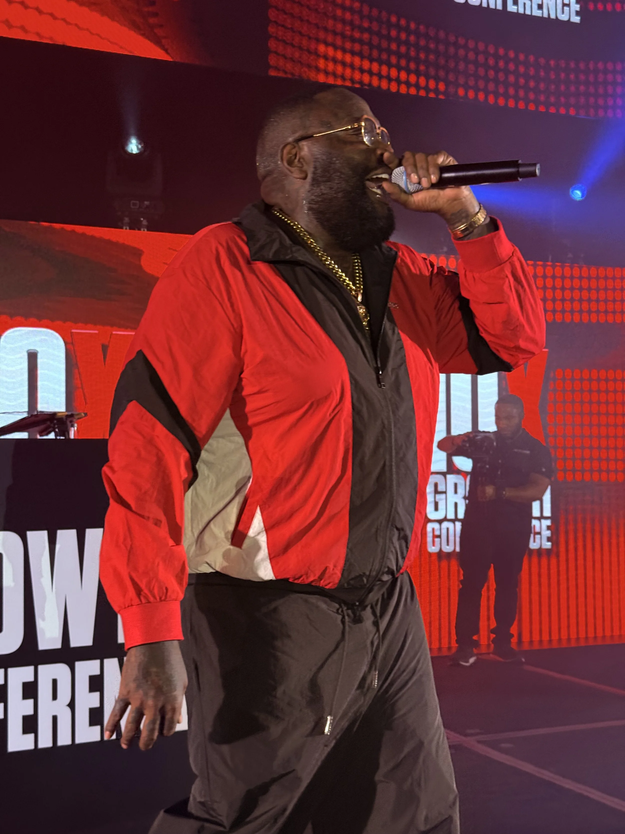 A man with glasses, a beard, and gold jewelry singing into a microphone on stage during a concert. He's wearing a red and black jacket and black pants. In the background, a DJ or sound technician is visible in front of a large digital screen with red