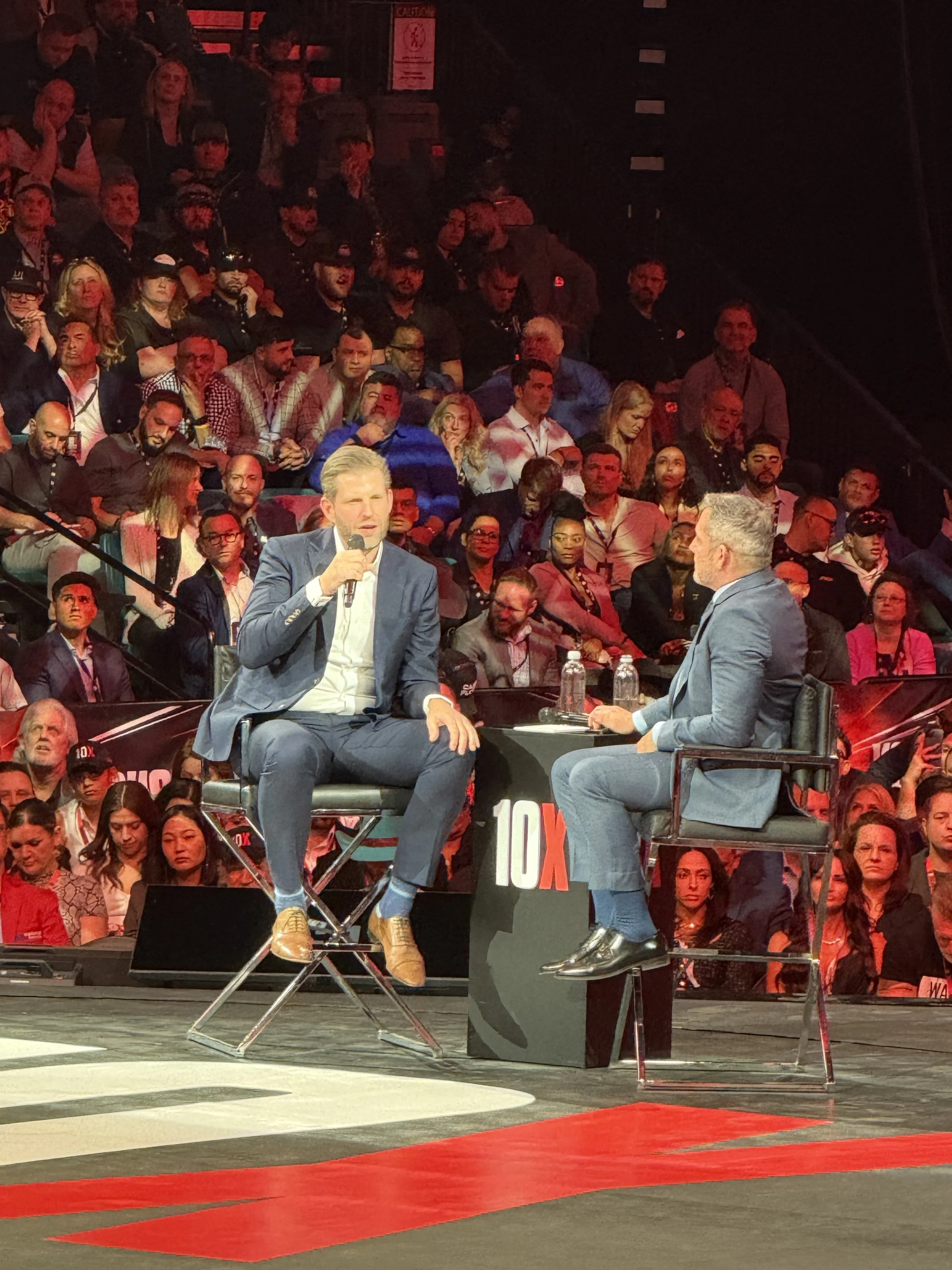 Two men in suits having a conversation during a live event on a stage, with a large audience in the background and the '10X' logo on a table between them.