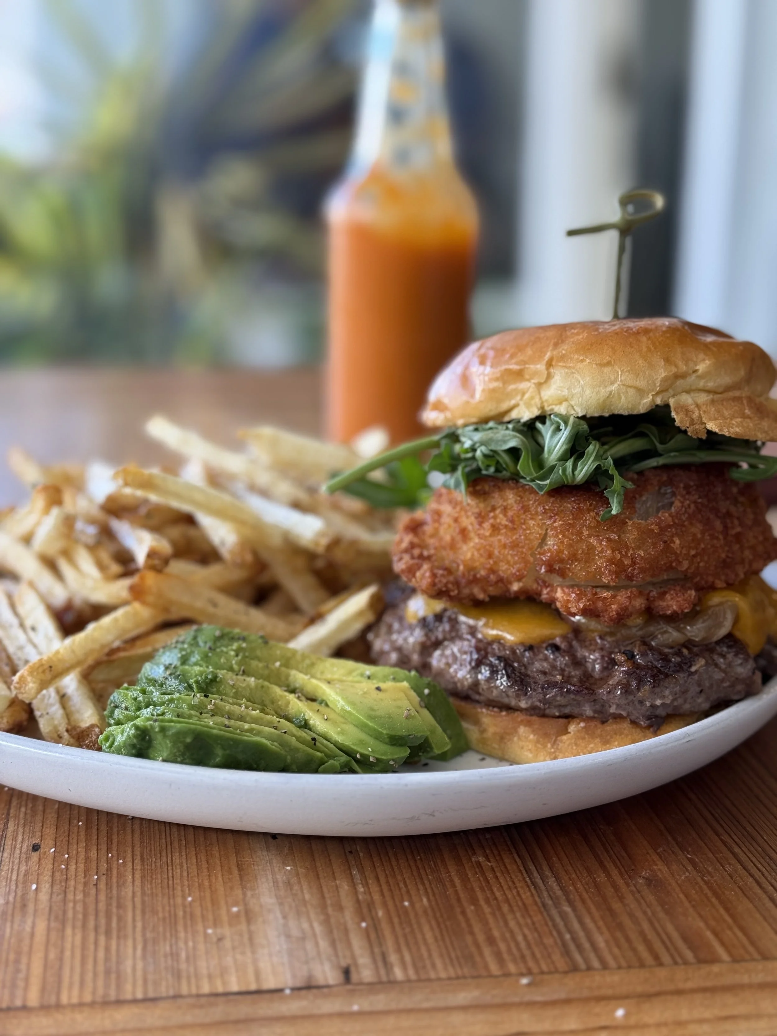 A plate with a cheeseburger, French fries, and sliced avocado. In the background, there is a bottle of hot sauce.