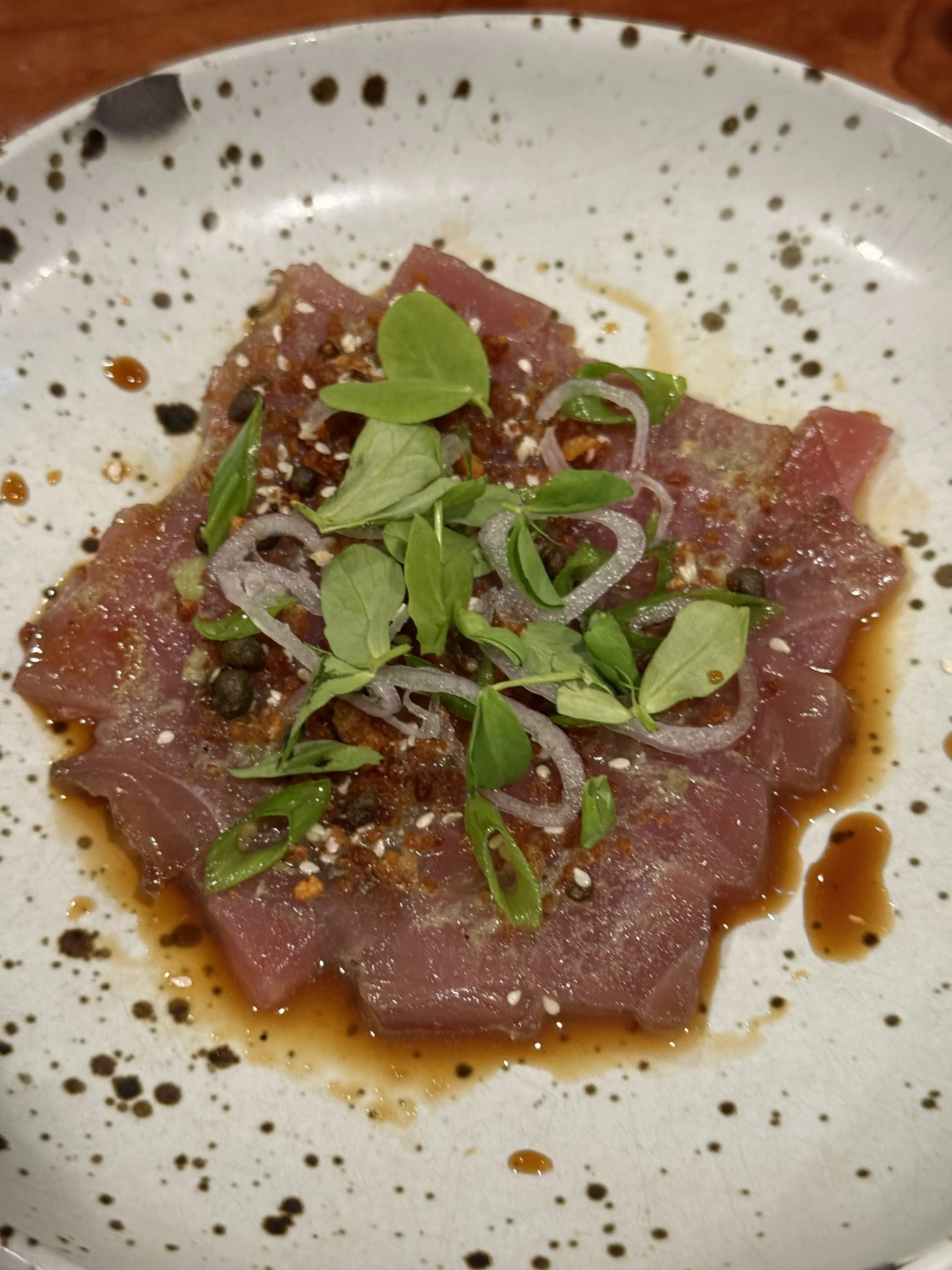 Raw tuna sashimi topped with microgreens, sliced onions, black pepper, and sauce on a white, speckled plate.