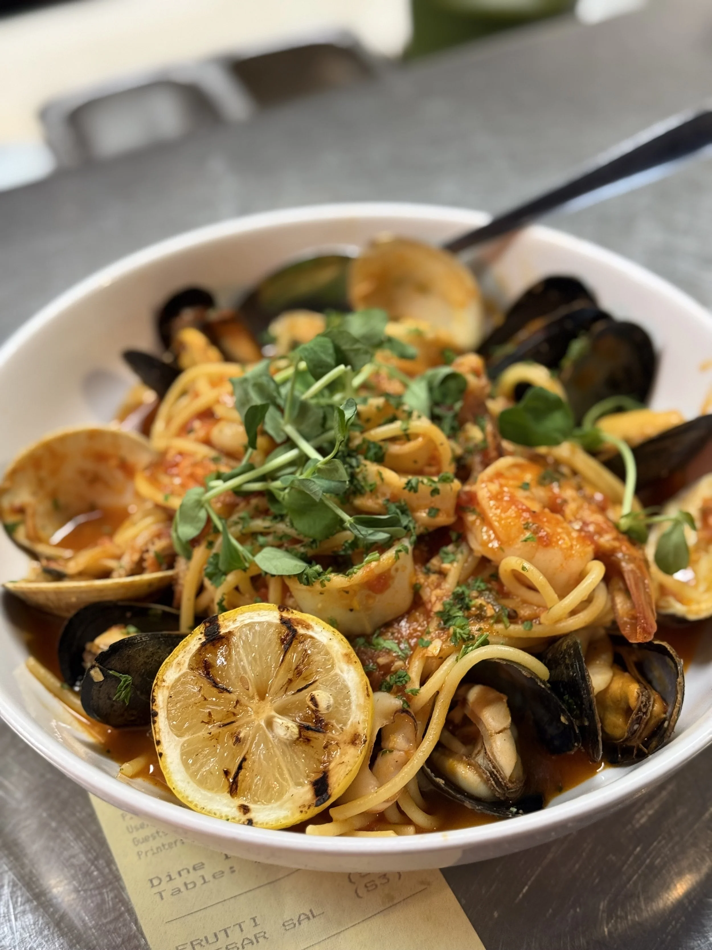 Seafood pasta with grilled lemon slice garnished with herbs, served in a white bowl on a gray surface.