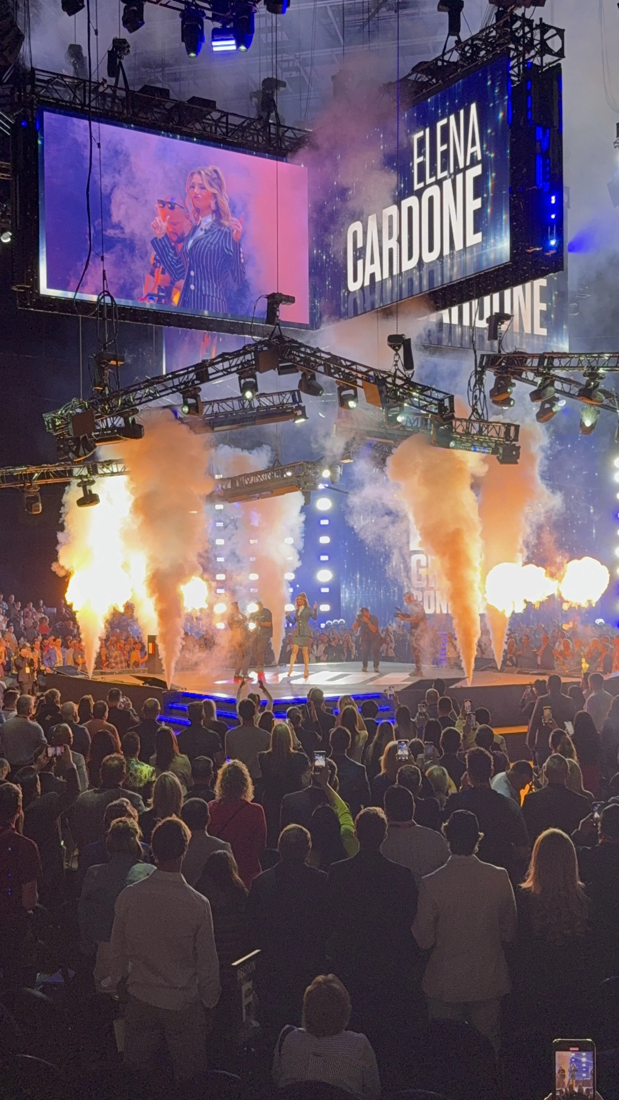 Elena Cardone performing on stage at a concert or event, with pyrotechnics and large screens displaying her name and image in the background, with a packed audience watching.