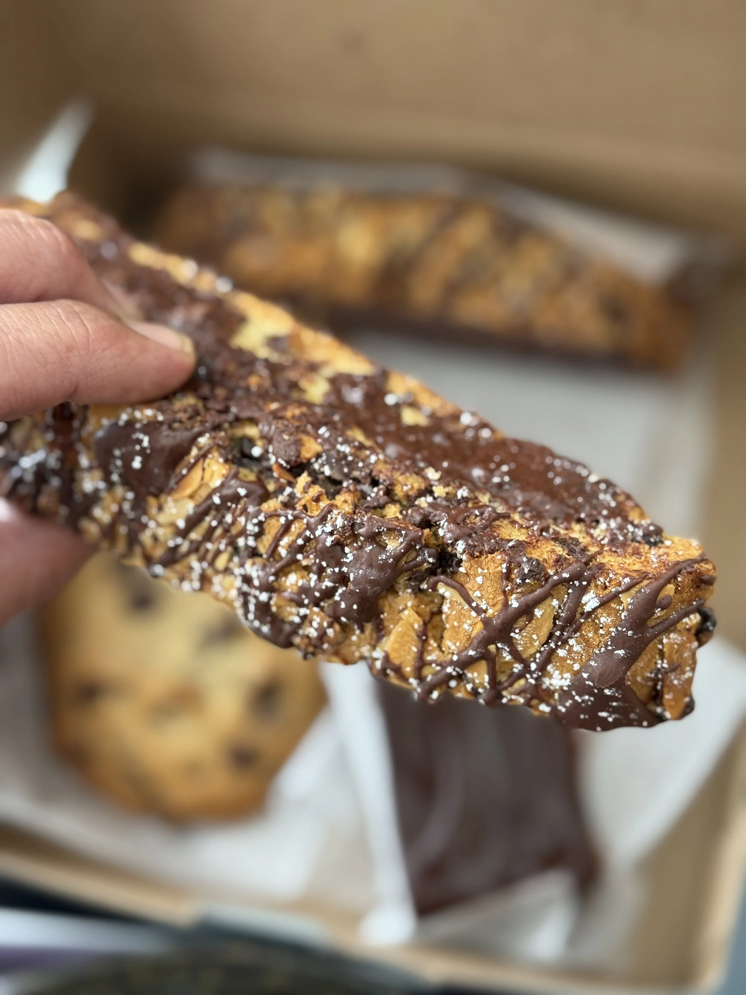 Close-up of a chocolate chip cookie bar drizzled with chocolate and sprinkled with powdered sugar, held by a person's hand.