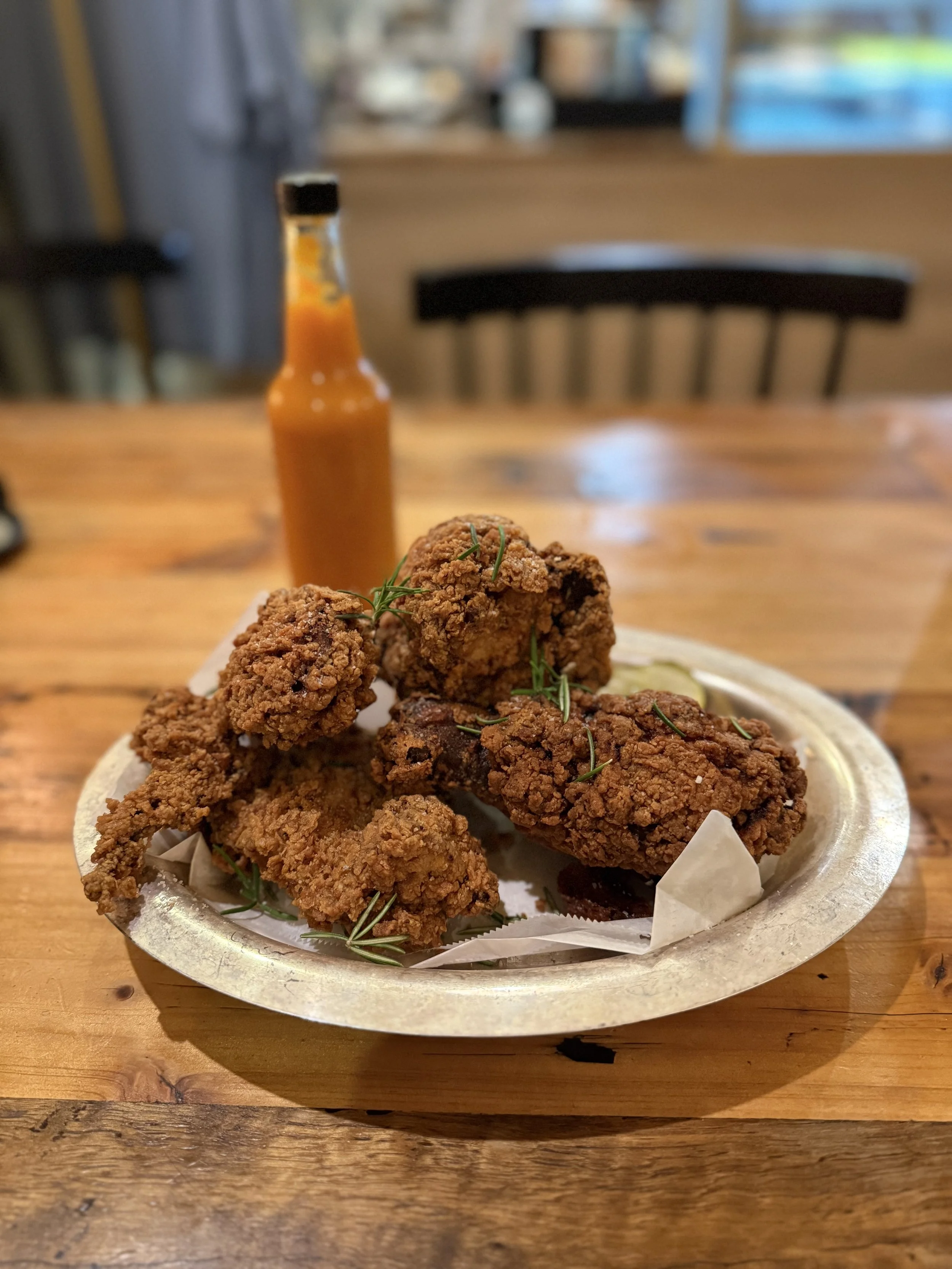 A plate of fried chicken pieces garnished with herbs with a bottle of hot sauce in the background on a wooden table.