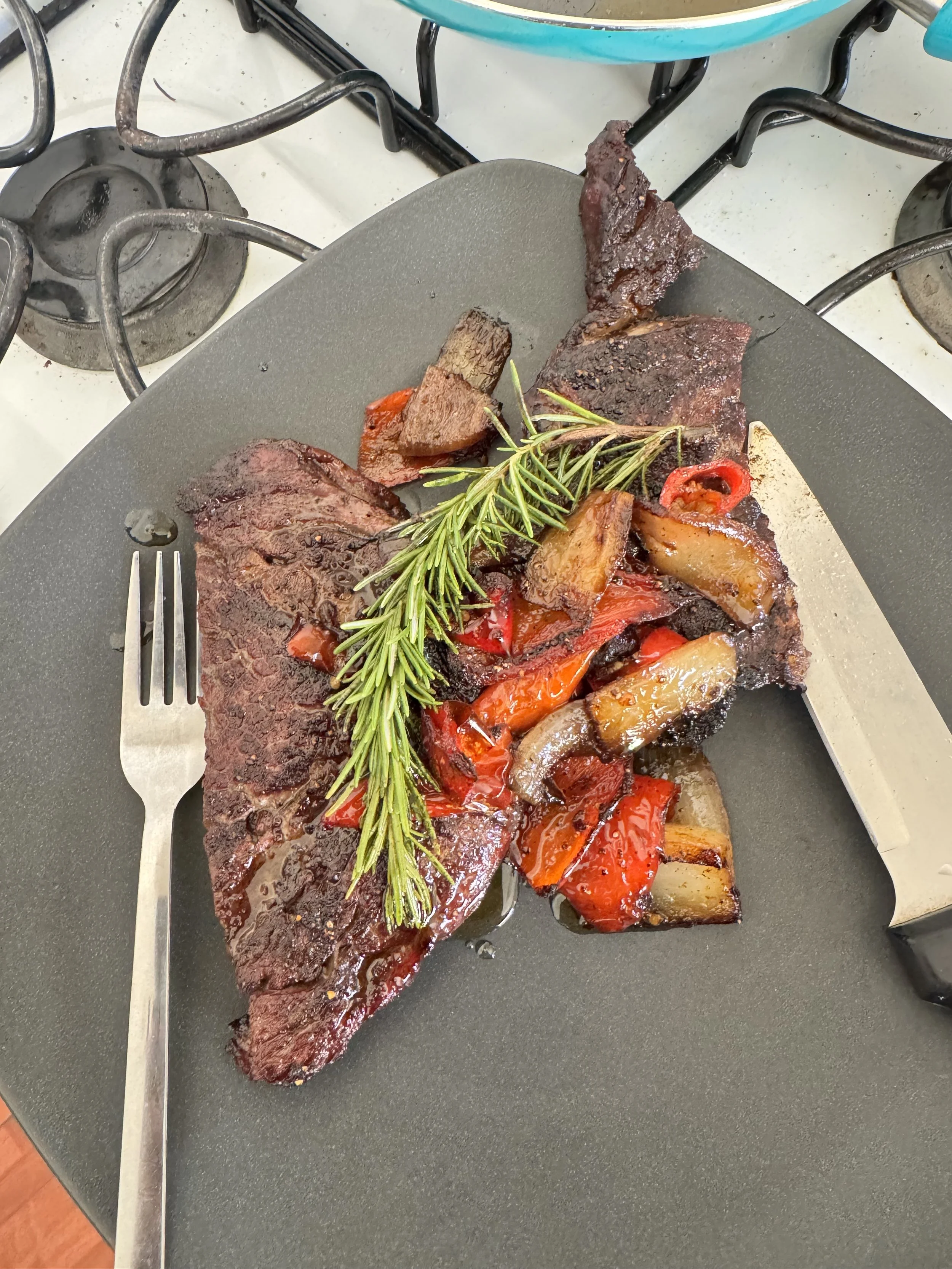 Cooked beef steak garnished with fresh rosemary, served with roasted red peppers and onions on a black plate.