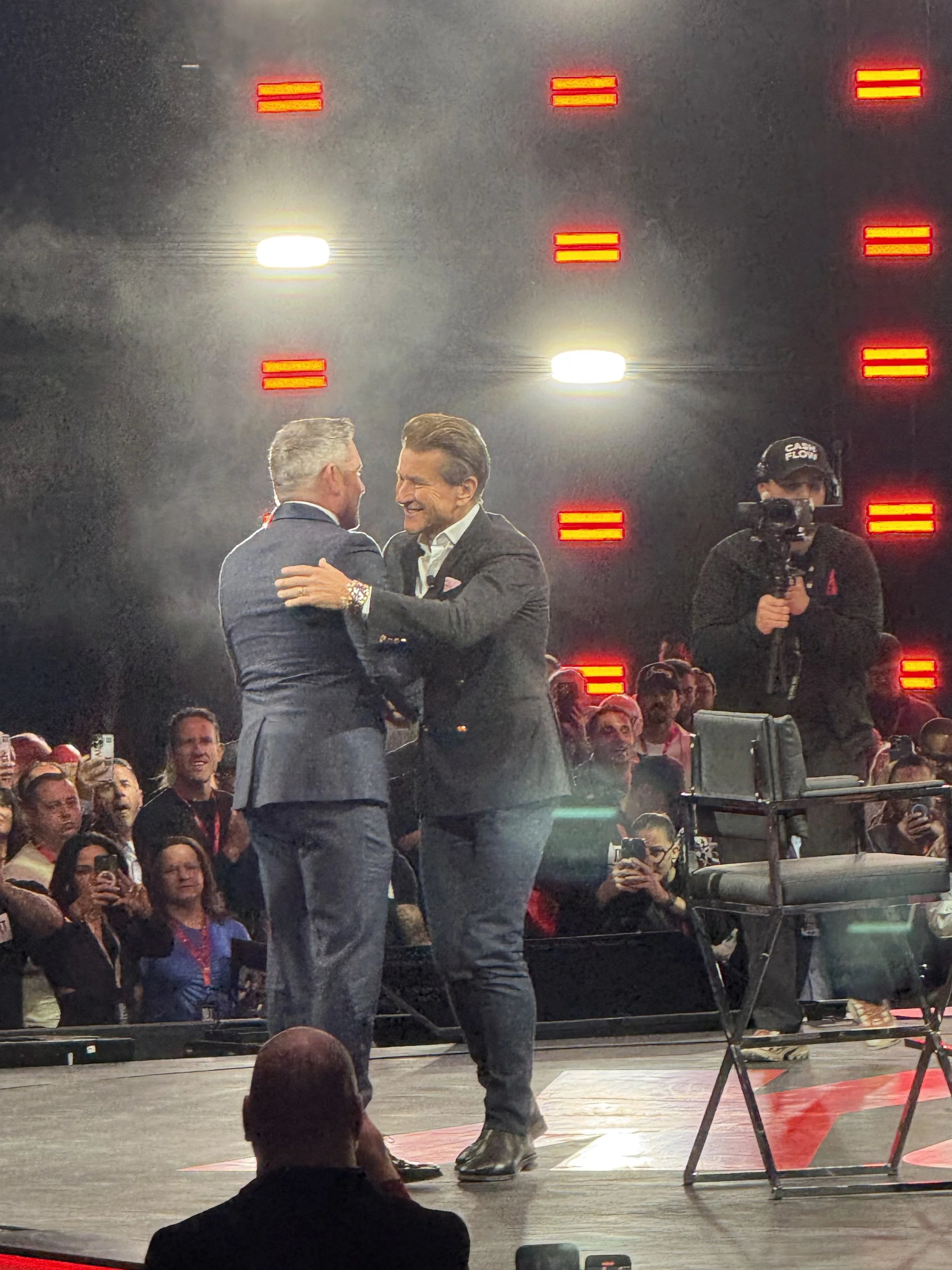 Two men in suits are greeting each other warmly on stage, surrounded by a crowd of spectators. A cameraman is capturing the moment, and bright stage lights are in the background.