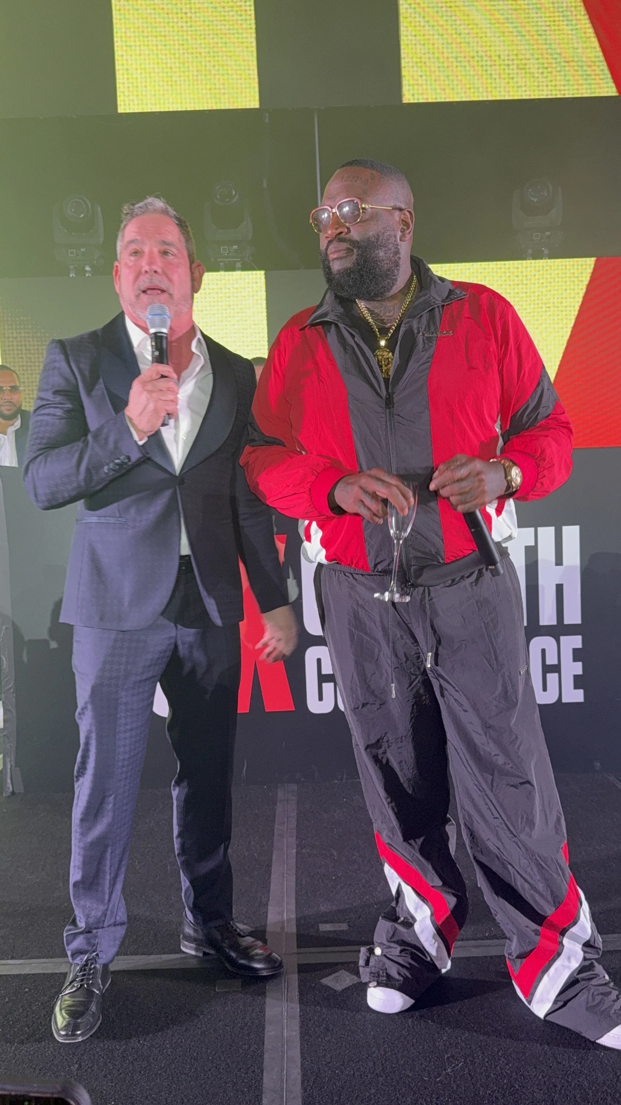 Two men on stage at a formal event, one in a dark gray suit holding a microphone, the other in a red and black track suit holding a champagne glass, with LED screens in the background.