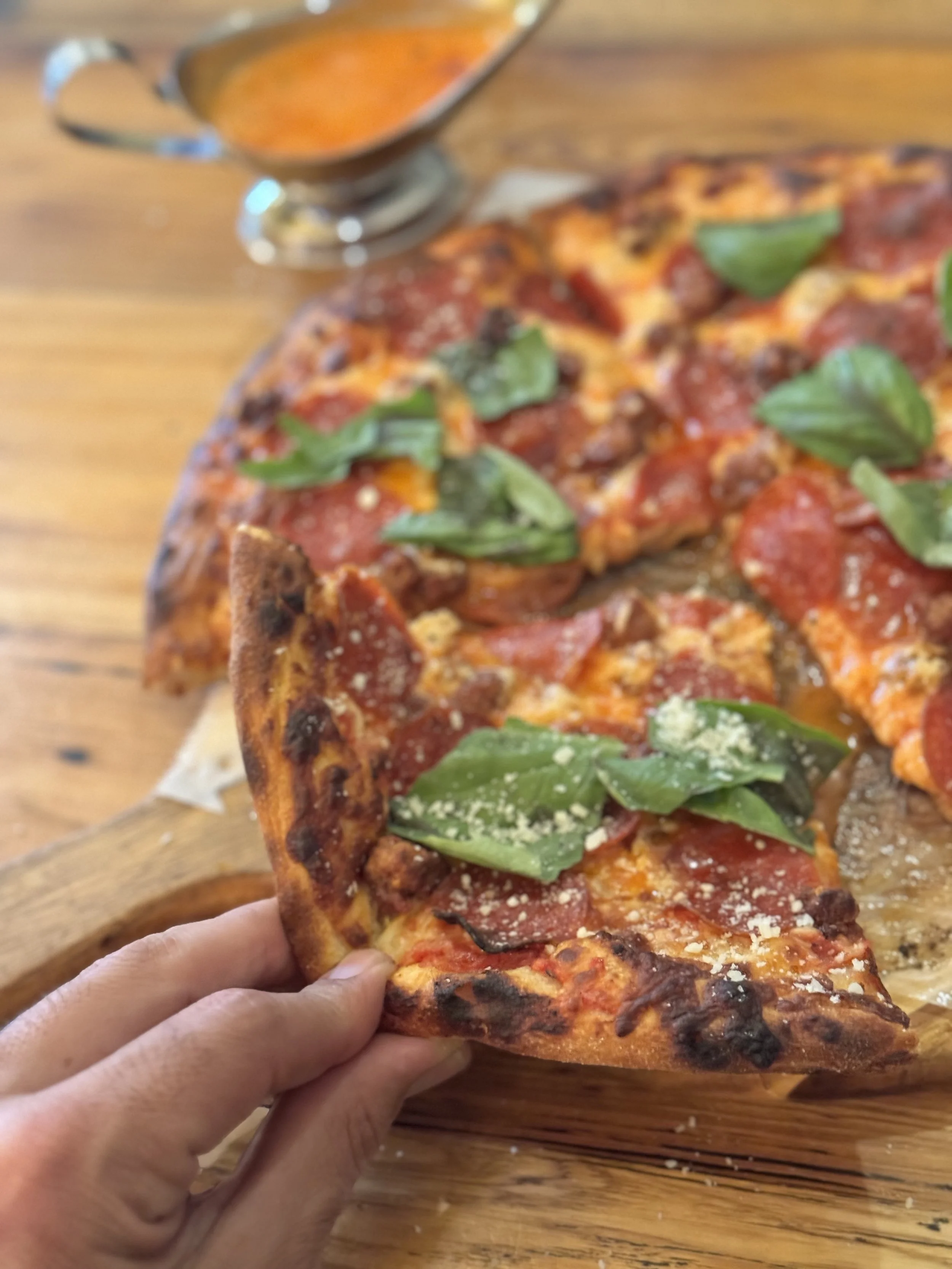 Slice of pepperoni pizza with basil leaves and grated cheese on a wooden pizza peel, with a bowl of orange sauce or dressing in the background.