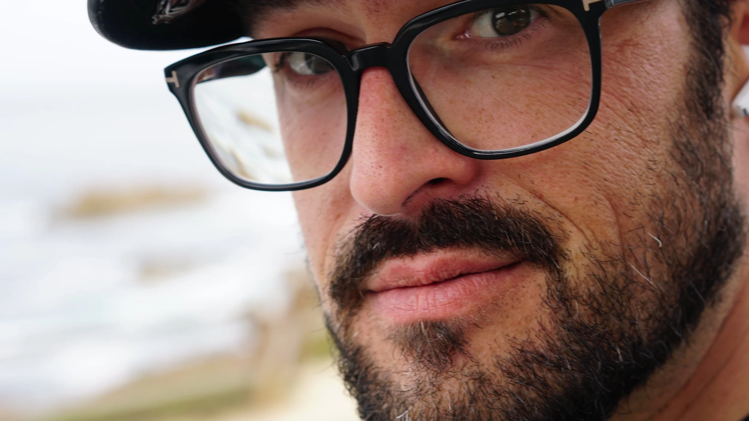 Close-up of a man with glasses and a beard, outdoors with a cloudy sky in the background.