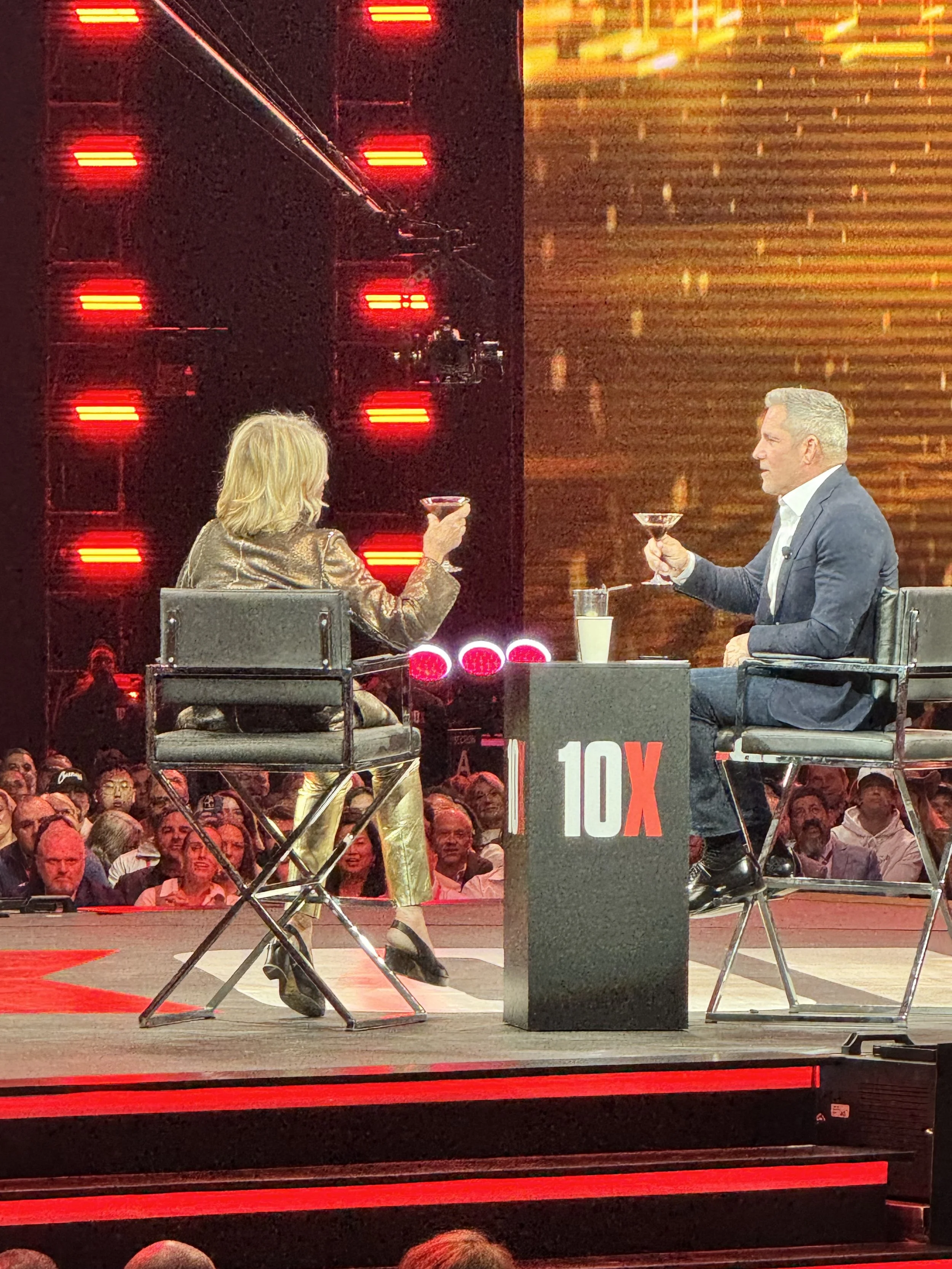 TV interview or talk show scene with two hosts sitting on stage, facing each other, clinking glasses, with a crowd in the background and red stage lights.
