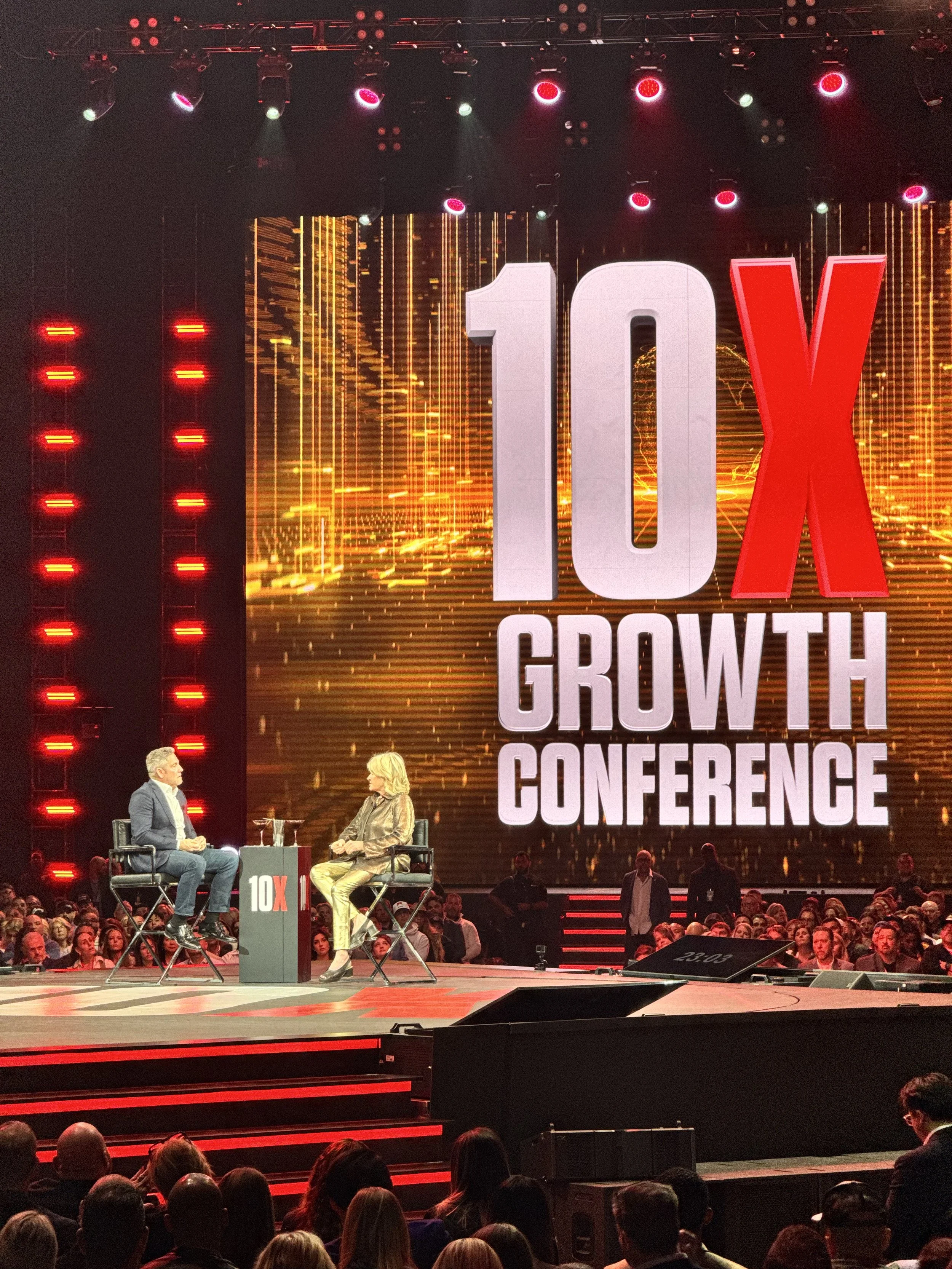 A stage at the 10X Growth Conference showing two people sitting on chairs engaged in conversation, with a large screen behind them displaying the conference logo and the words '10X Growth Conference.' The stage is illuminated with red and white light