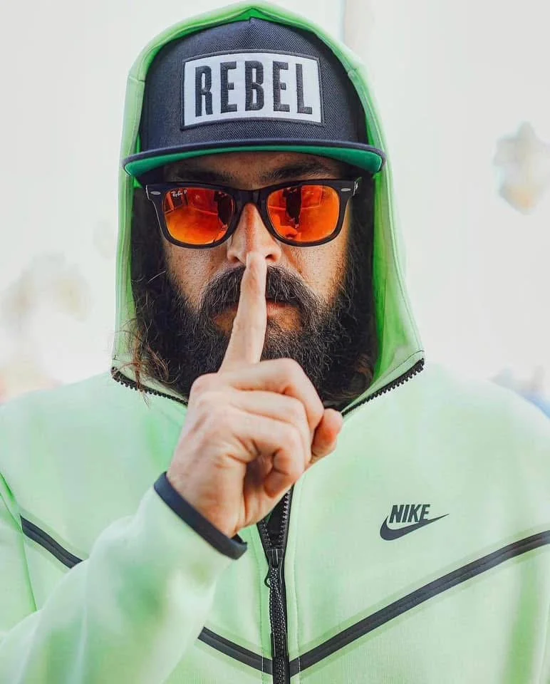 A man wearing a black cap with the word 'REBEL' on it, orange reflective sunglasses, a neon green Nike jacket with the hood up, and making a 'shush' gesture with his index finger over his lips.