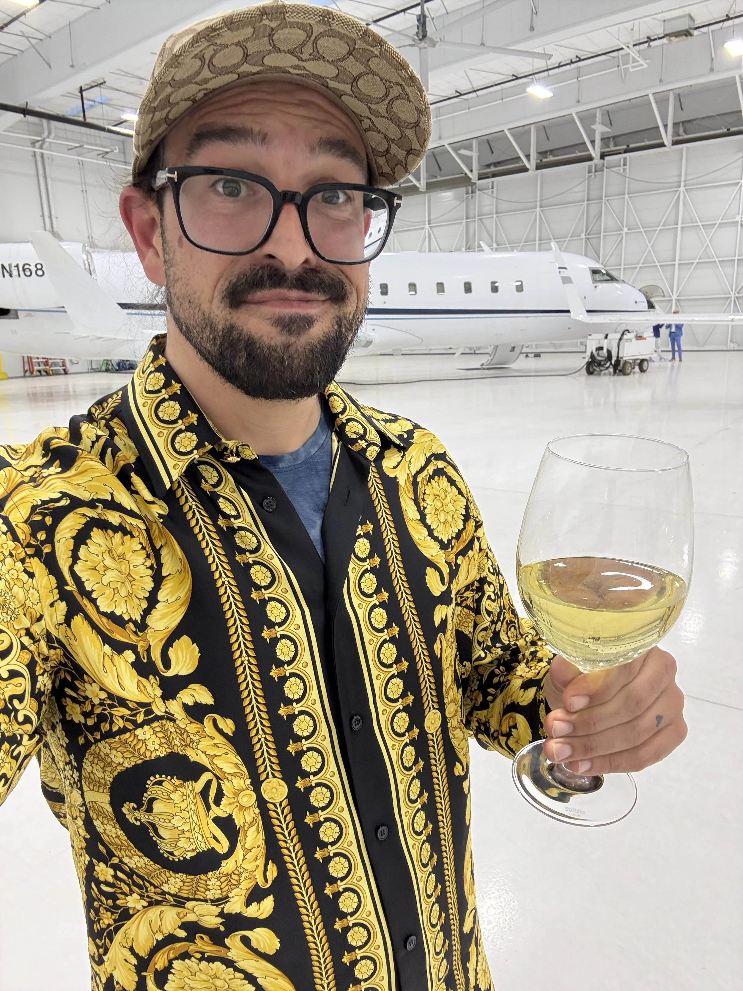 A man with glasses, a beard, and a patterned hat holding a glass of white wine inside an airplane hangar with a private jet in the background.