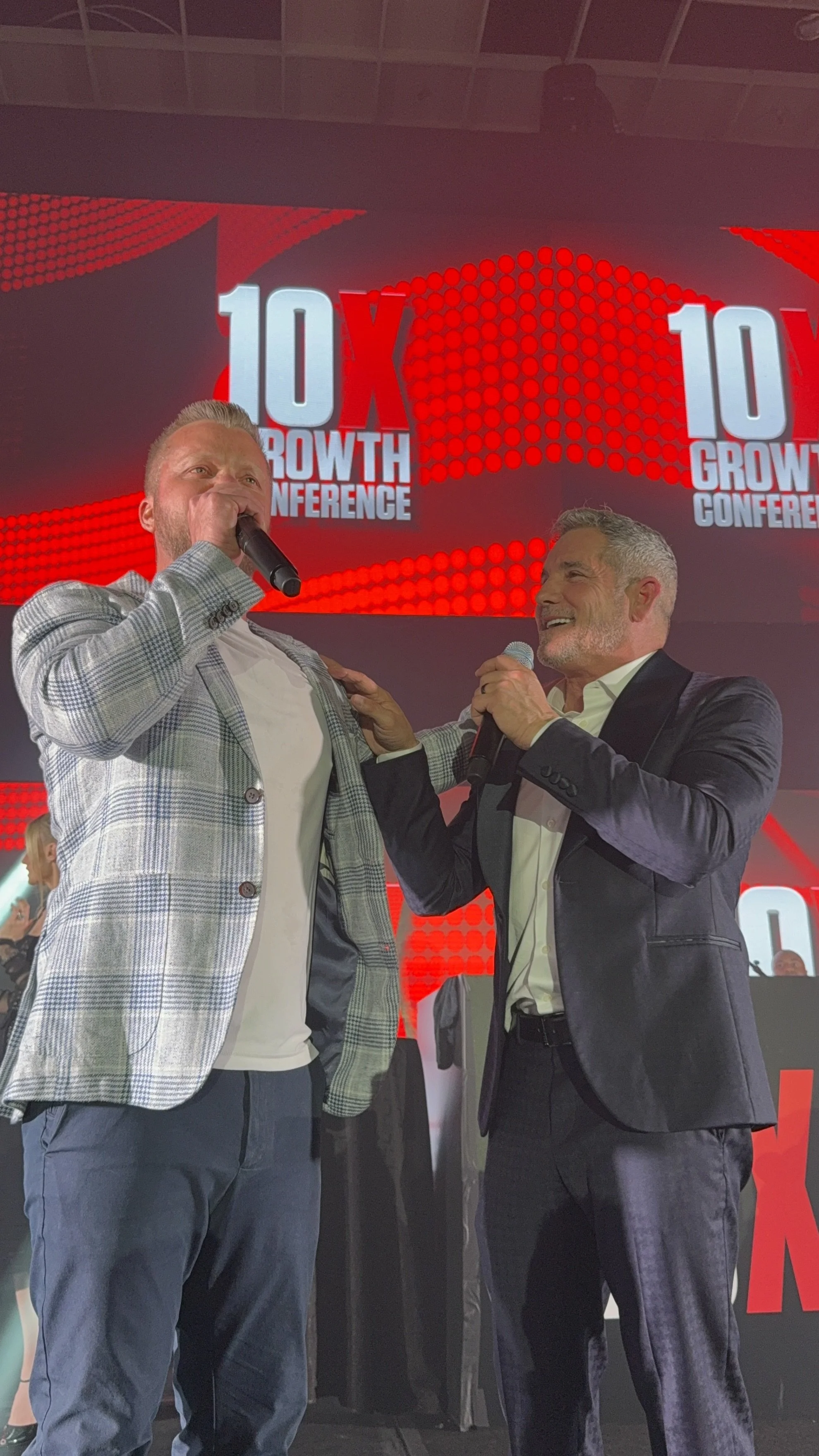 Two men are on stage at the 10X Growth Conference, holding microphones and engaging in a conversation or performance, with a large digital screen displaying the conference name in the background.