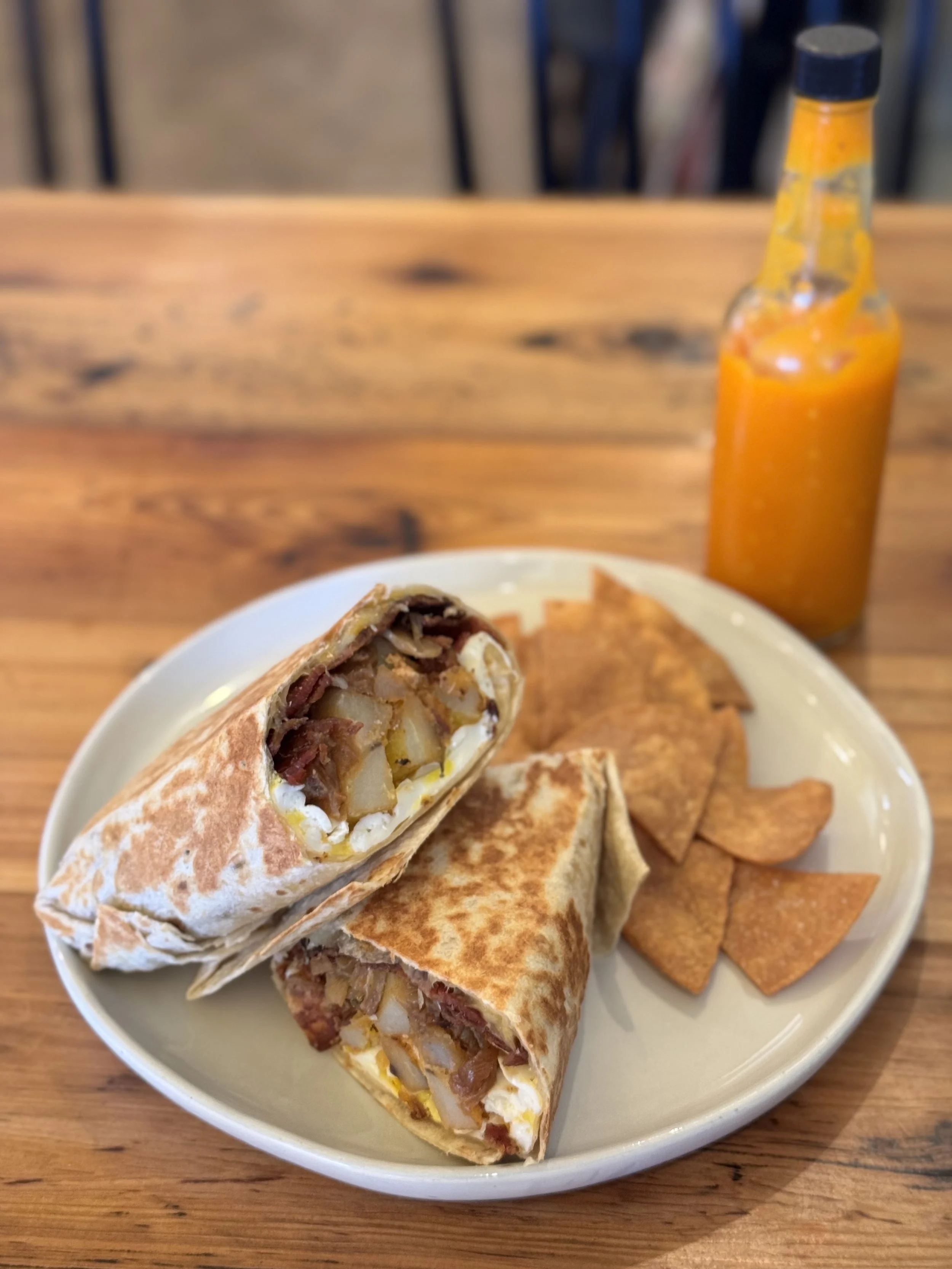 Plate with a beef, potato, and cheese burrito, sliced in half, with nacho chips and a bottle of hot sauce on a wooden table.