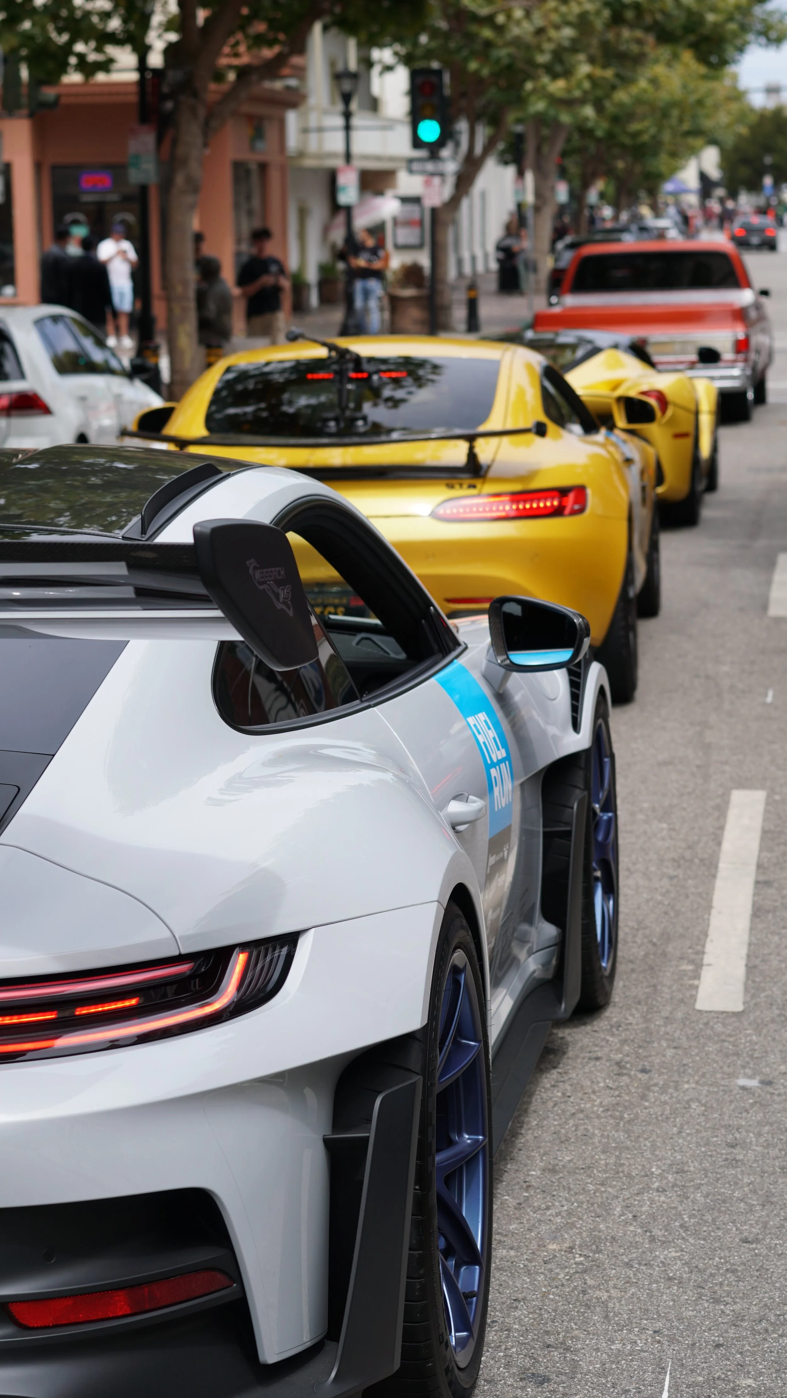 Several parked sports cars, including a white and black race car with a "Fuel Run" sticker, a yellow sports car, and a red sports car, on an urban street with trees, buildings, pedestrians, and traffic lights.